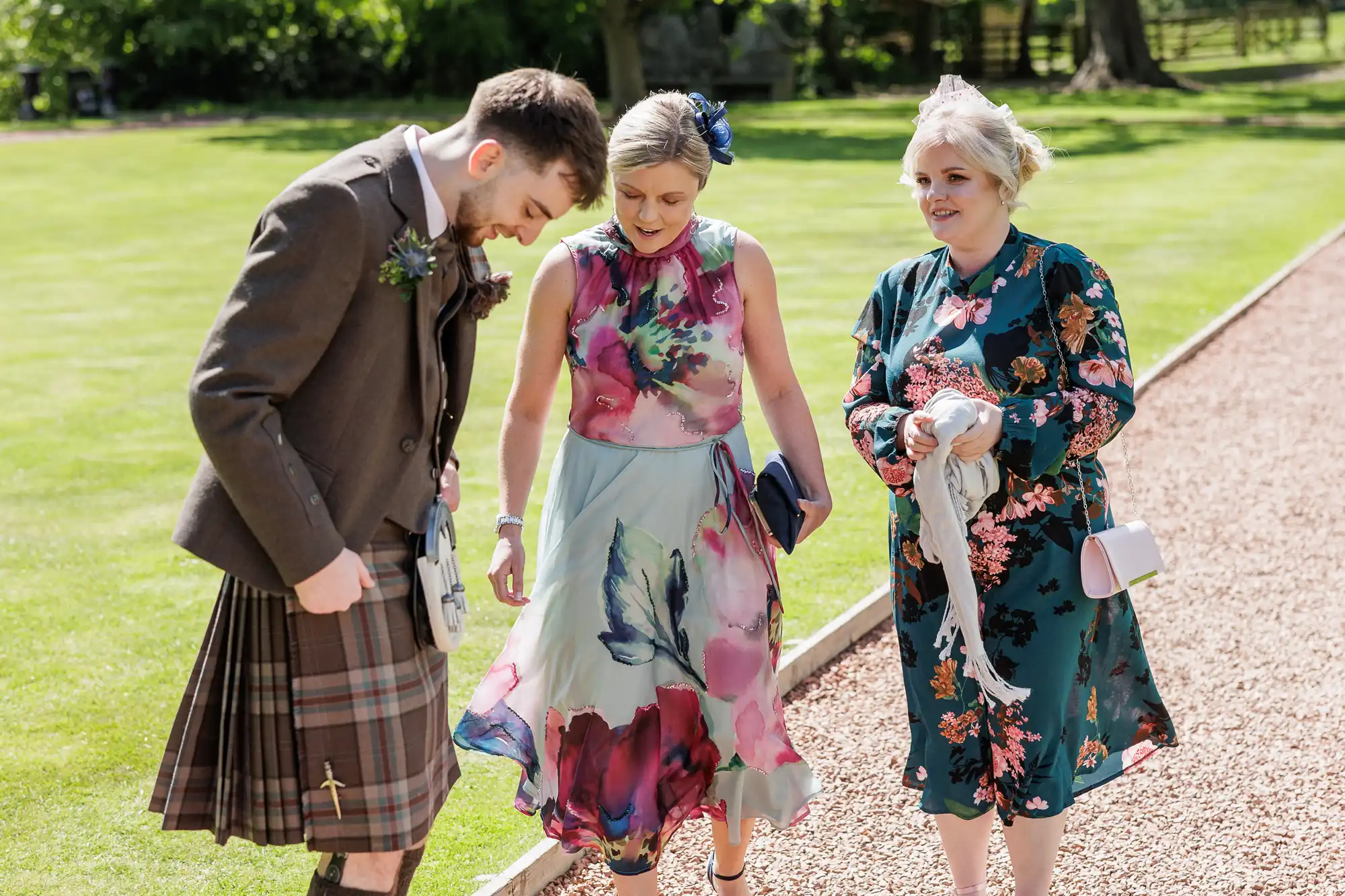Three people walk outdoors on a gravel path. The man on the left wears a brown jacket and tartan kilt. The woman in the middle wears a sleeveless floral dress, and the woman on the right wears a long-sleeved floral dress and holds a pale shawl. Green grass is in the background.