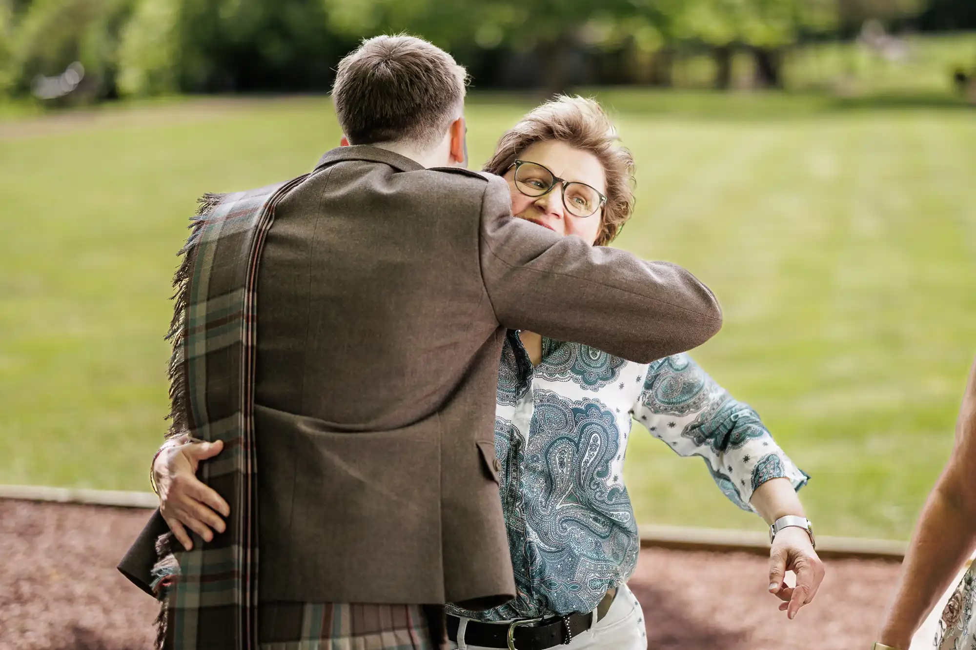 A man in a brown jacket hugs a smiling woman wearing glasses and a patterned shirt, outdoors on a sunny day in a grassy park. The woman appears happy, and both are positioned near a path, with trees and grass in the background.