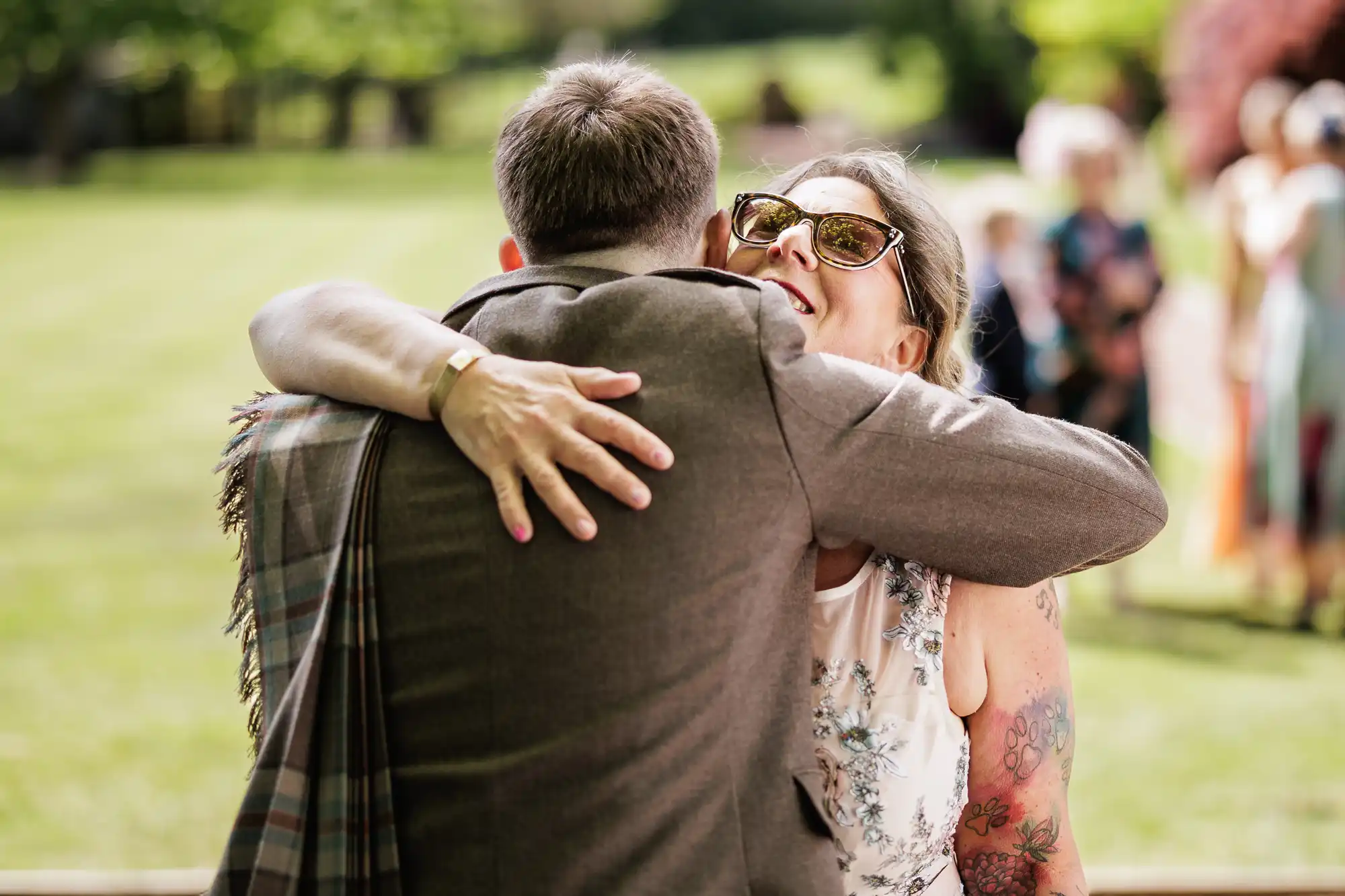 A man in a brown suit embraces a smiling woman wearing sunglasses and a sleeveless floral top. Her arm, showing colourful tattoos, is visible. They are outdoors on a sunny day with blurred greenery and people in the background.
