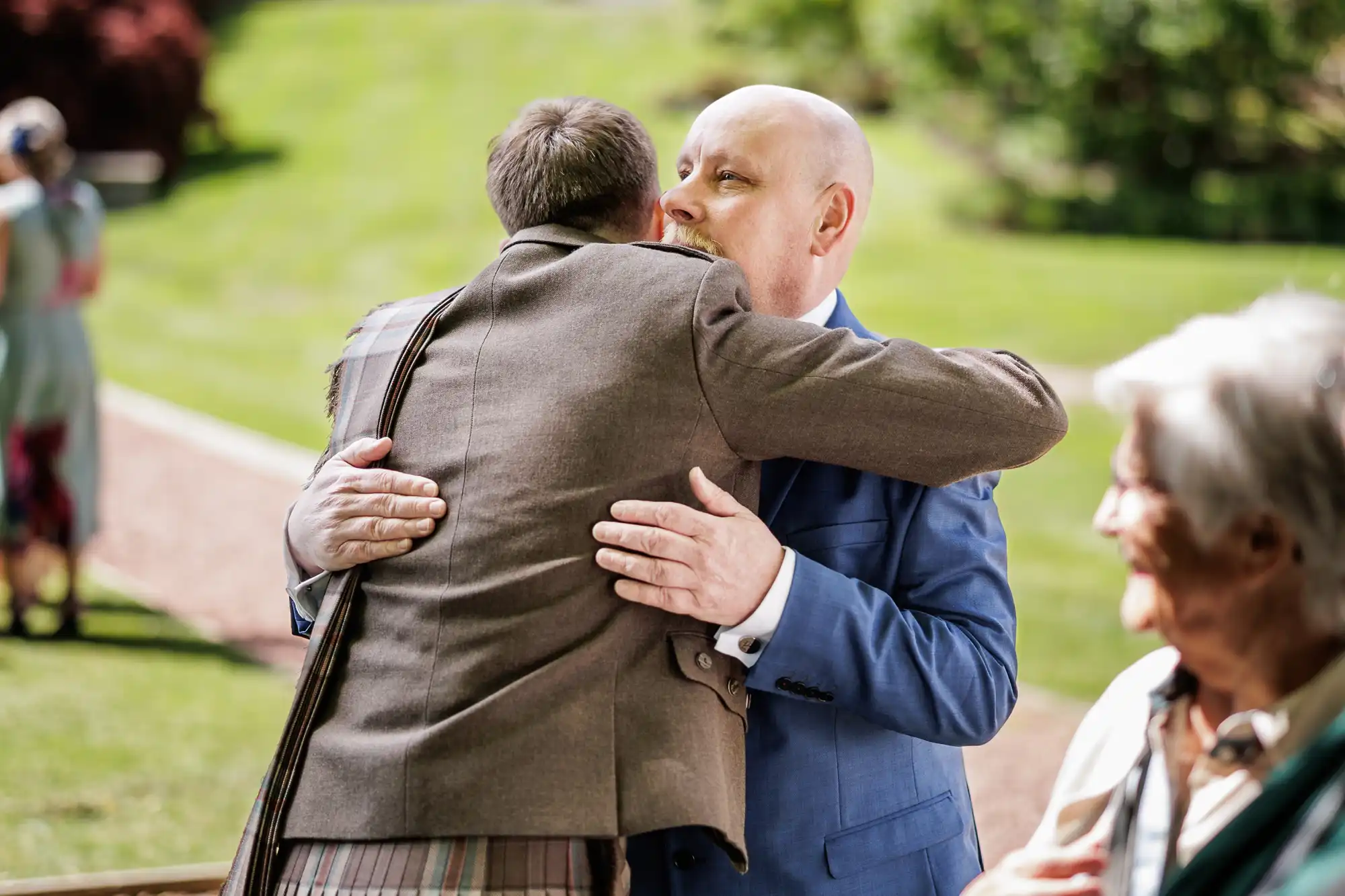 Two men embrace warmly outdoors; one wears a brown jacket and tartan kilt, the other a blue suit. Green lawn and trees are in the background. An elderly person with white hair and glasses stands nearby, partially out of focus.