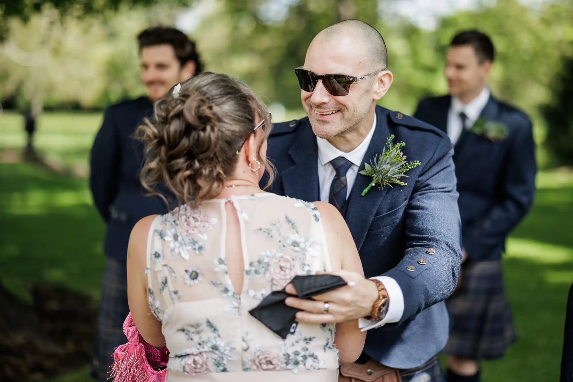 A smiling man in sunglasses and a navy jacket with a buttonhole greets a woman in a sheer floral dress outdoors. Two men in matching attire and kilts stand blurred in the background on a sunny, green lawn.