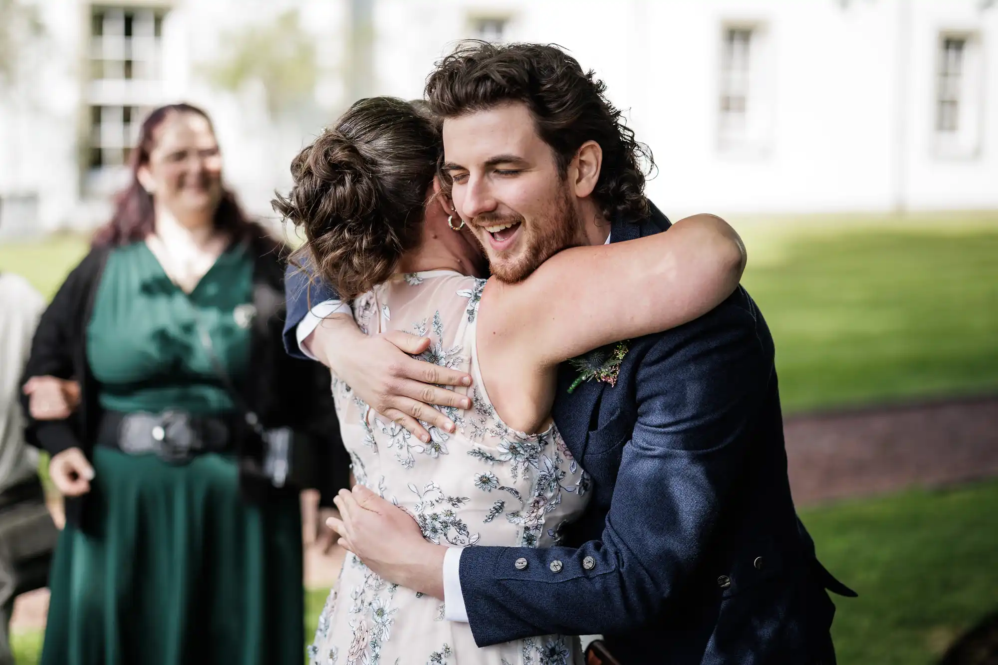 A man in a navy suit hugs a woman in a light floral dress outdoors, both smiling warmly. Another woman in a green dress stands in the blurred background on a grassy lawn, with a white building behind them. The scene feels joyful and celebratory.