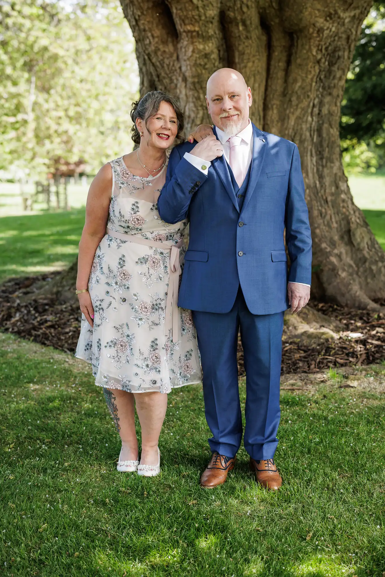 A smiling couple stands on grass in front of a large tree. The woman wears a sleeveless, knee-length floral dress and white shoes. The man is in a blue suit, light pink tie, and brown shoes. They appear happy and are posing closely together outdoors.