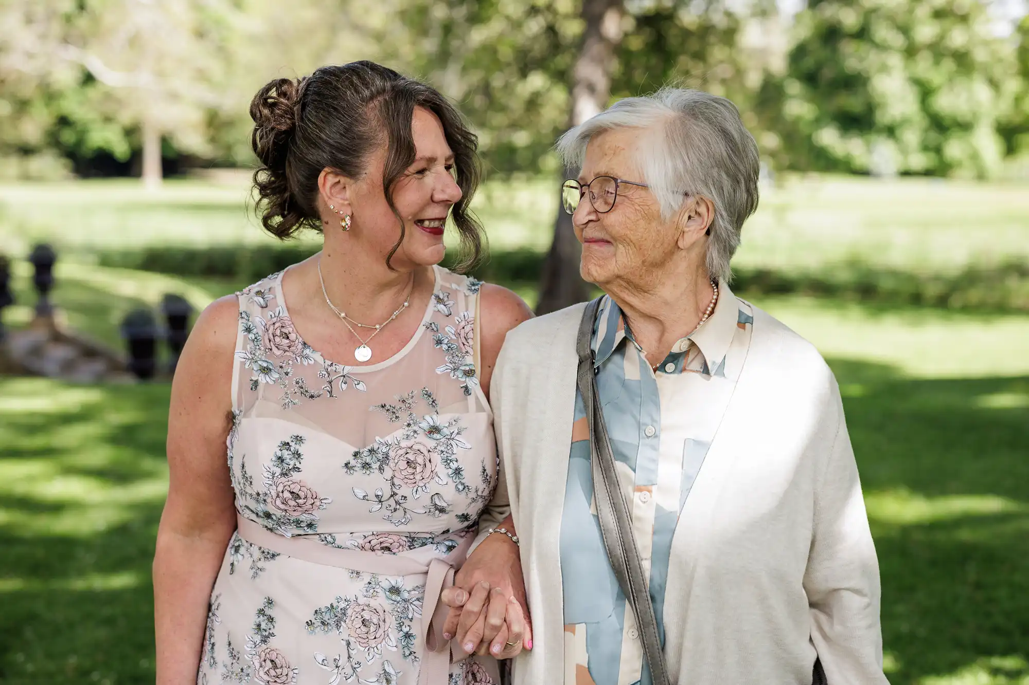 A smiling middle-aged woman in a floral sleeveless dress holds hands and walks with an elderly woman in glasses, a light cardigan, and blouse. They look at each other warmly, standing outside in a sunlit, green park.
