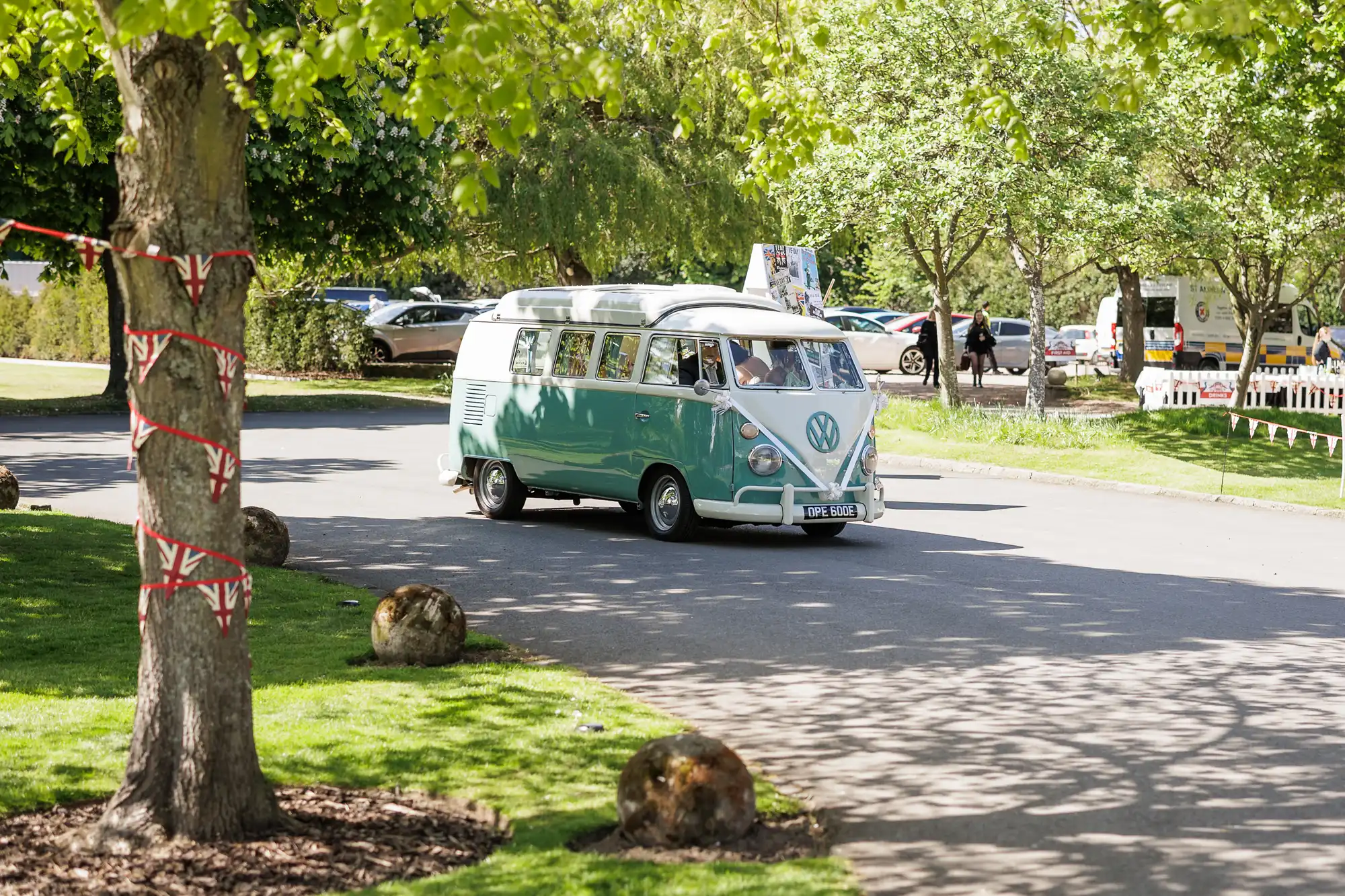 A vintage light blue and white Volkswagen camper van drives on a sunny, tree-lined road in a park. The trees cast dappled shadows, and parked cars are visible in the background. The grass is lush and green, with large stones scattered near the road.