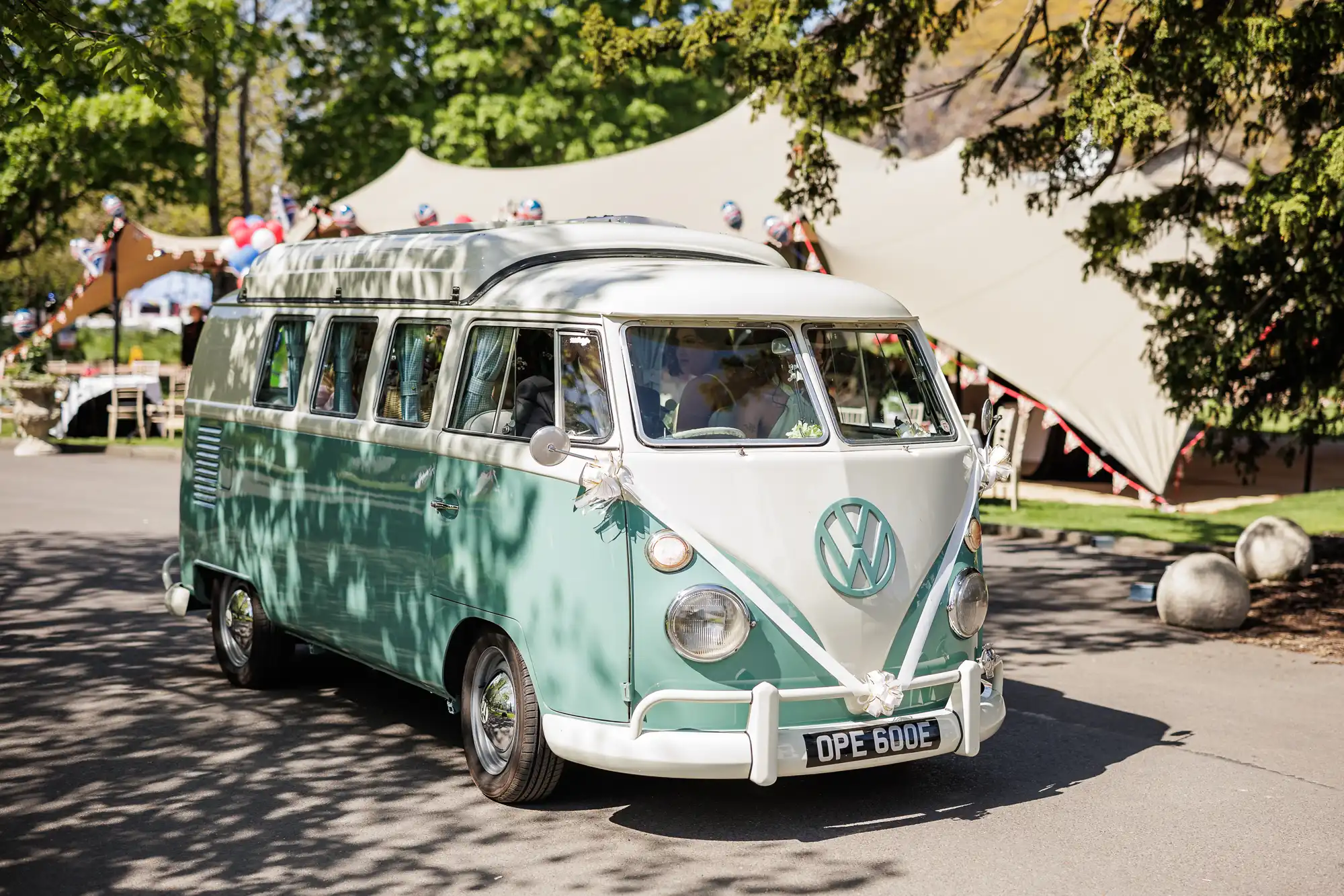 A vintage mint green and white Volkswagen camper van decorated with white ribbons drives on a sunny day. Trees and a large marquee with bunting are in the background, suggesting a festive outdoor event, possibly a wedding or celebration.