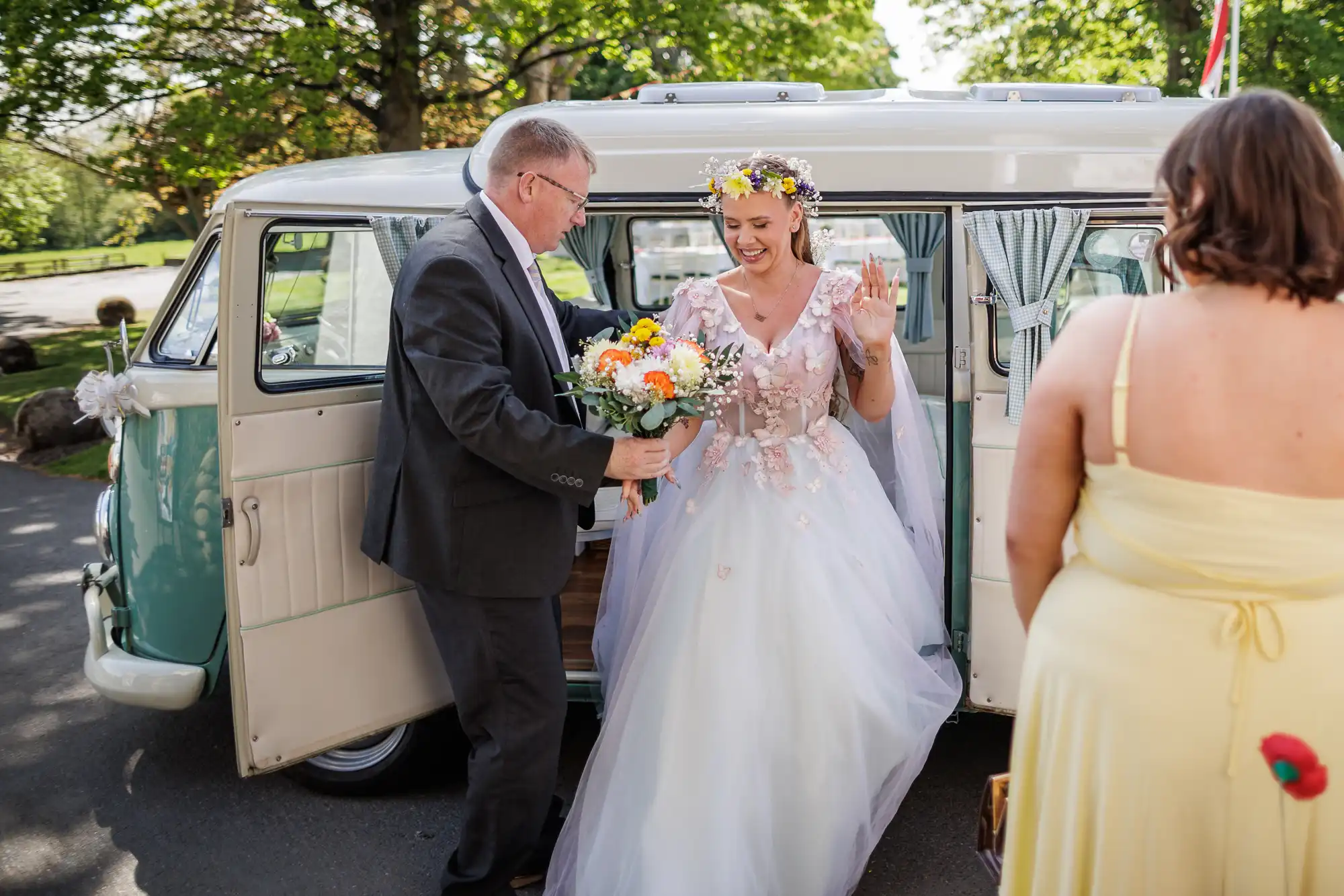 A smiling bride in a flower crown and white gown steps out of a vintage van, holding a bouquet. A man in a suit offers his hand to help her. A woman in a yellow dress stands nearby. Trees and sunlight are visible in the background.