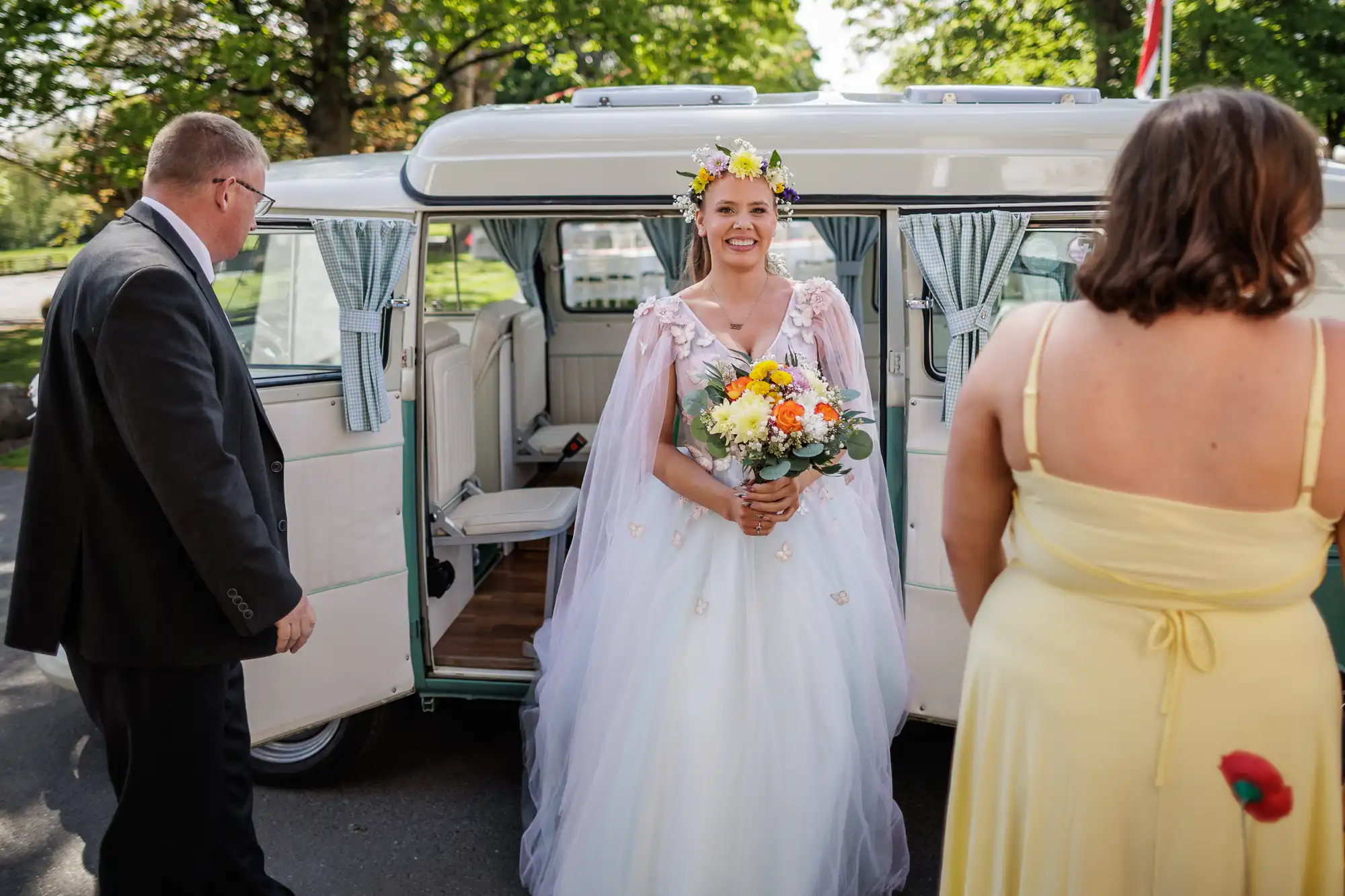 A smiling bride in a white gown and floral crown holds a bouquet of yellow and white flowers, standing by a vintage van with open doors. Two people, one in a suit and one in a yellow dress, face her. Trees and sunlight fill the background.