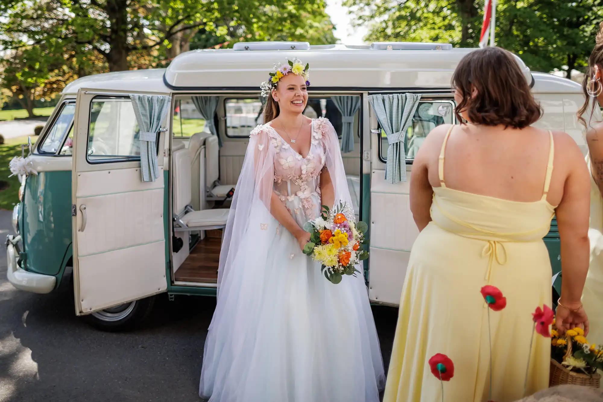 A bride in a white dress and flower crown smiles, holding a colourful bouquet, standing beside a vintage van with open doors. Another woman in a yellow dress stands with her back to the camera. Green trees and sunlight fill the background.