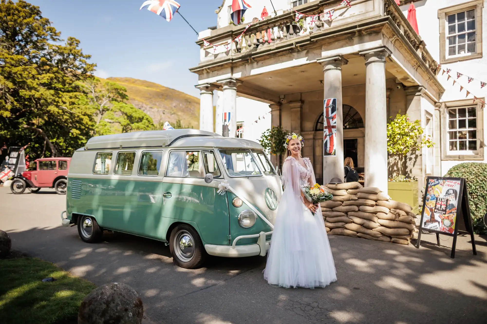 A bride in a white gown and flower crown stands beside a vintage green VW camper van outside a grand building decorated with flags, bunting, and sandbags. She holds a bouquet and smiles, with sunlight and trees in the background.
