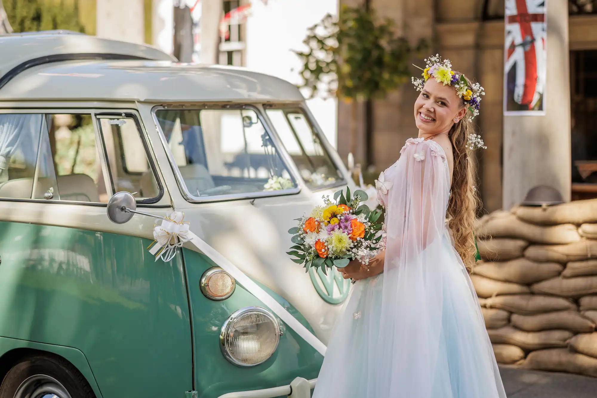 A smiling woman in a flowing white dress and floral crown holds a bouquet of colourful flowers beside a vintage green and white van decorated with ribbons. She stands outdoors on a sunny day, with Union Jack flags and sandbags in the background.