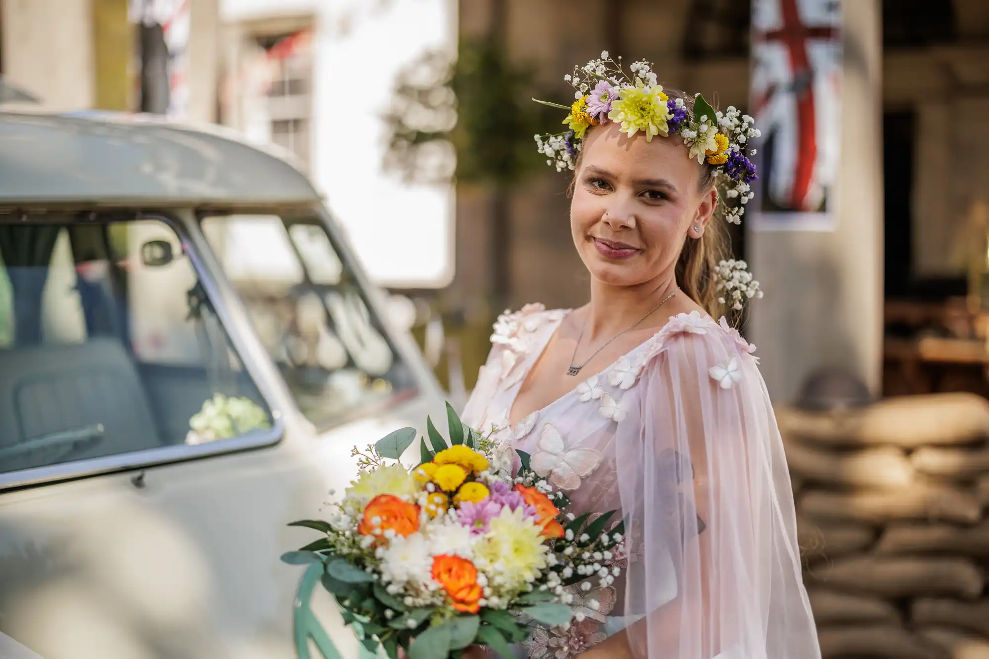 A woman in a pale pink dress with flower appliqués and a colourful floral crown smiles whilst holding a bouquet of flowers. She stands beside a vintage car. In the background are blurred flags and stacked sacks, giving a festive, rustic vibe.
