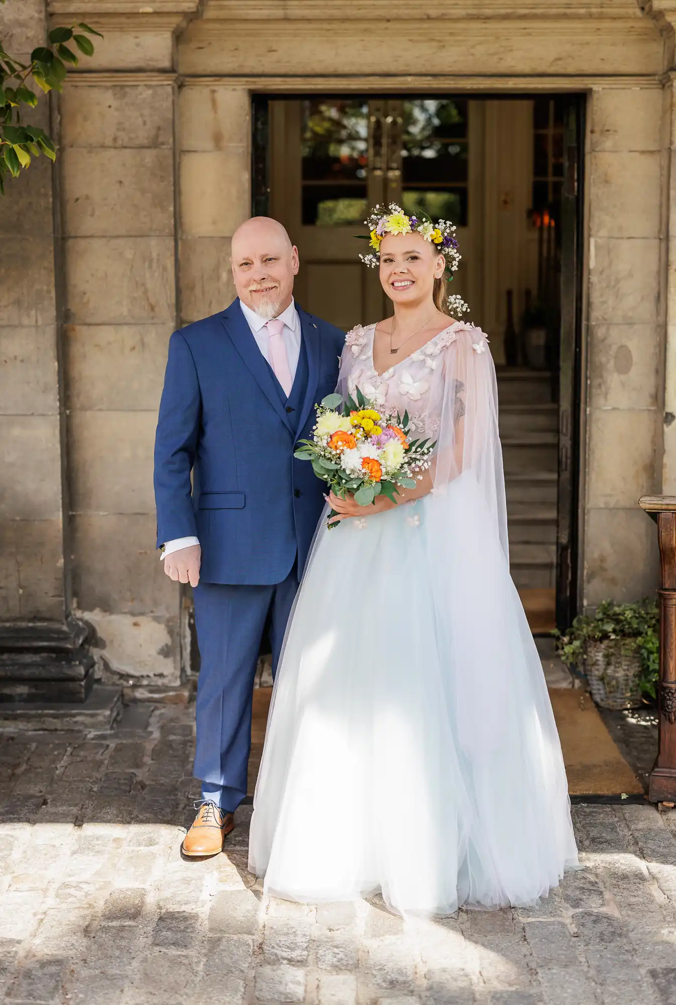 A smiling couple stands outside a stone building. The man wears a blue suit and pink tie. The woman wears a light blue gown with sheer sleeves, a floral crown, and holds a bouquet of colourful flowers. Sunlight brightens the scene.