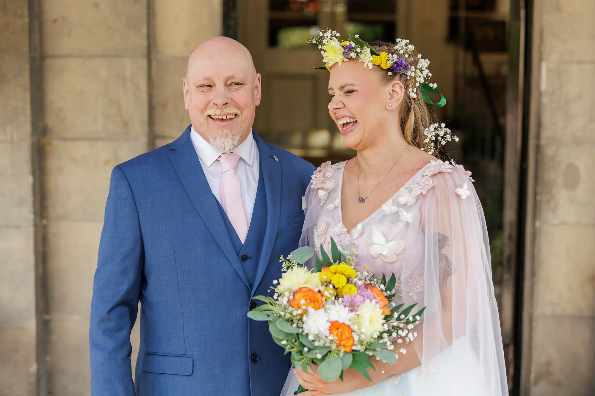 A smiling man in a blue suit and light pink tie stands next to a laughing woman in a floral dress and flower crown, holding a colourful bouquet with yellow, white, and orange flowers. They are outside, in front of a stone wall and doorway.