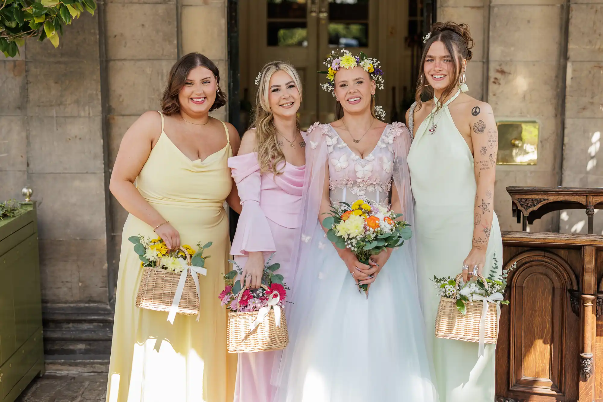 Four women stand smiling together outdoors. The bride is in a white gown with floral details and a flower crown, holding a bouquet. Her three bridesmaids wear pastel dresses in yellow, pink, and mint, each holding a basket of flowers. They pose by a stone wall.