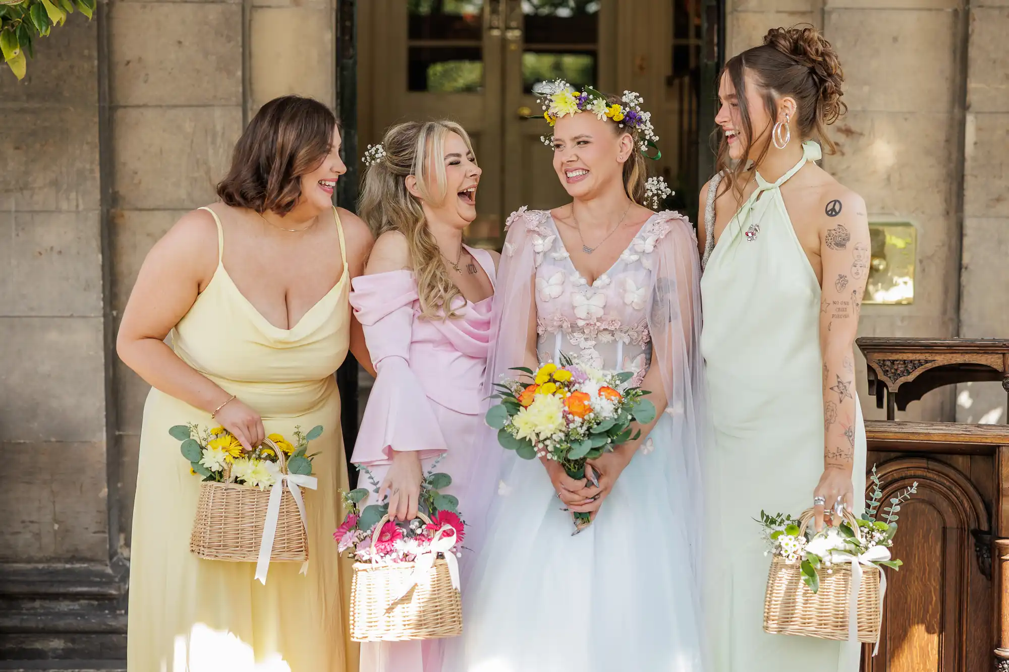 Four women in pastel dresses stand smiling together outdoors. The bride in a white gown and flower crown holds a bouquet; her three bridesmaids carry flower-filled baskets. They stand in front of a stone building, enjoying a joyful moment.