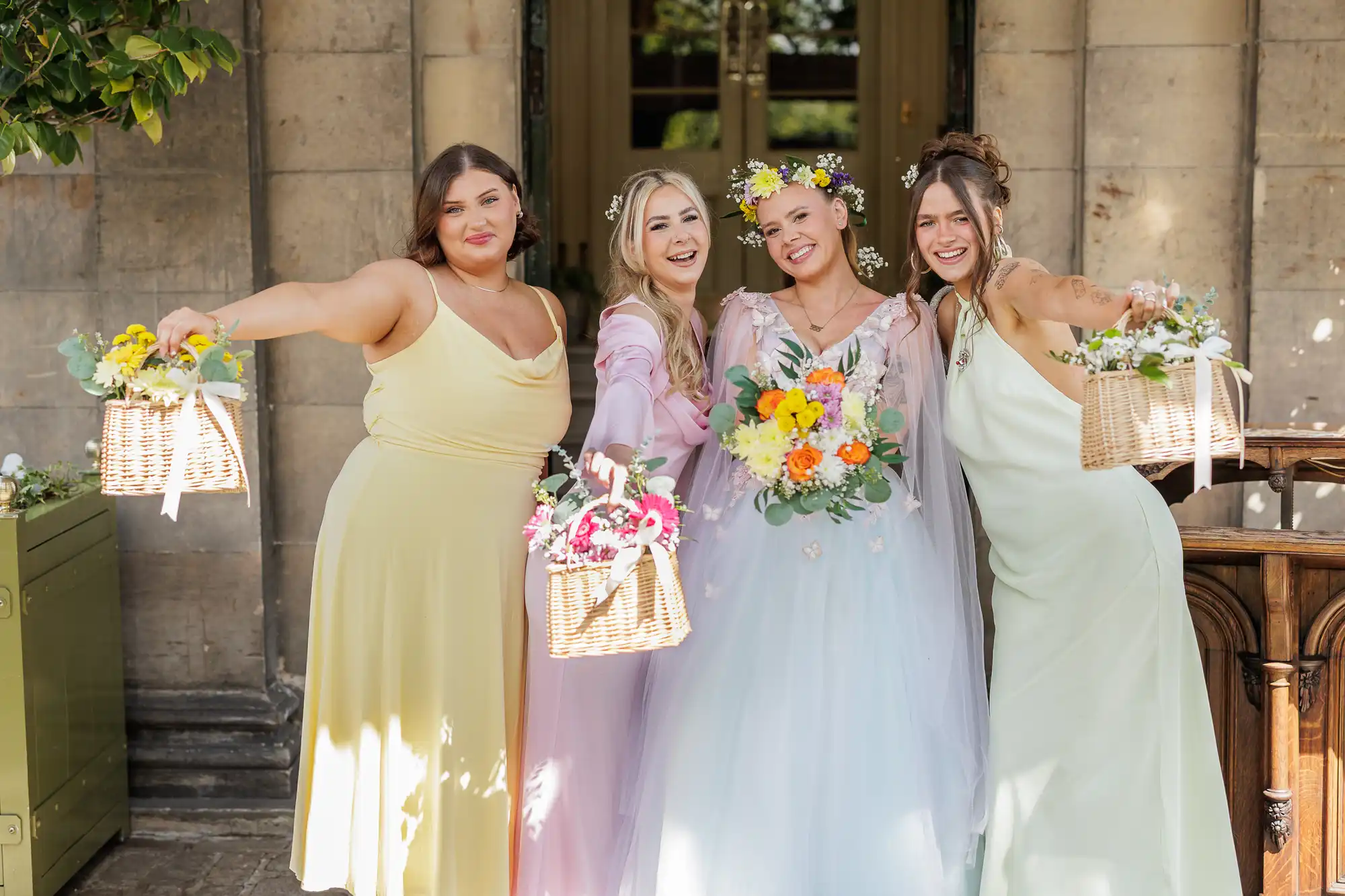 Four smiling women in pastel dresses stand outside a stone building, holding flower baskets out towards the camera. The central woman wears a white floral wedding dress and flower crown; the others wear yellow, pink, and pale green dresses, celebrating together.