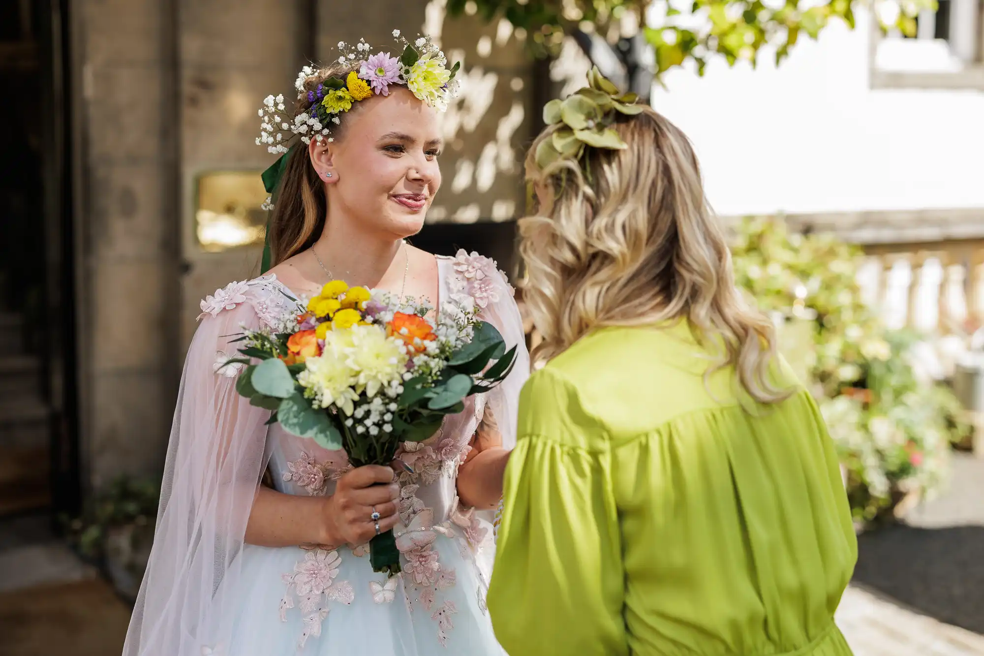 A smiling bride in a floral crown and blush pink dress holds a bouquet, chatting with a guest in a lime green dress and leafy headpiece, outdoors in bright sunlight. Lush greenery and part of a stone building are in the background.