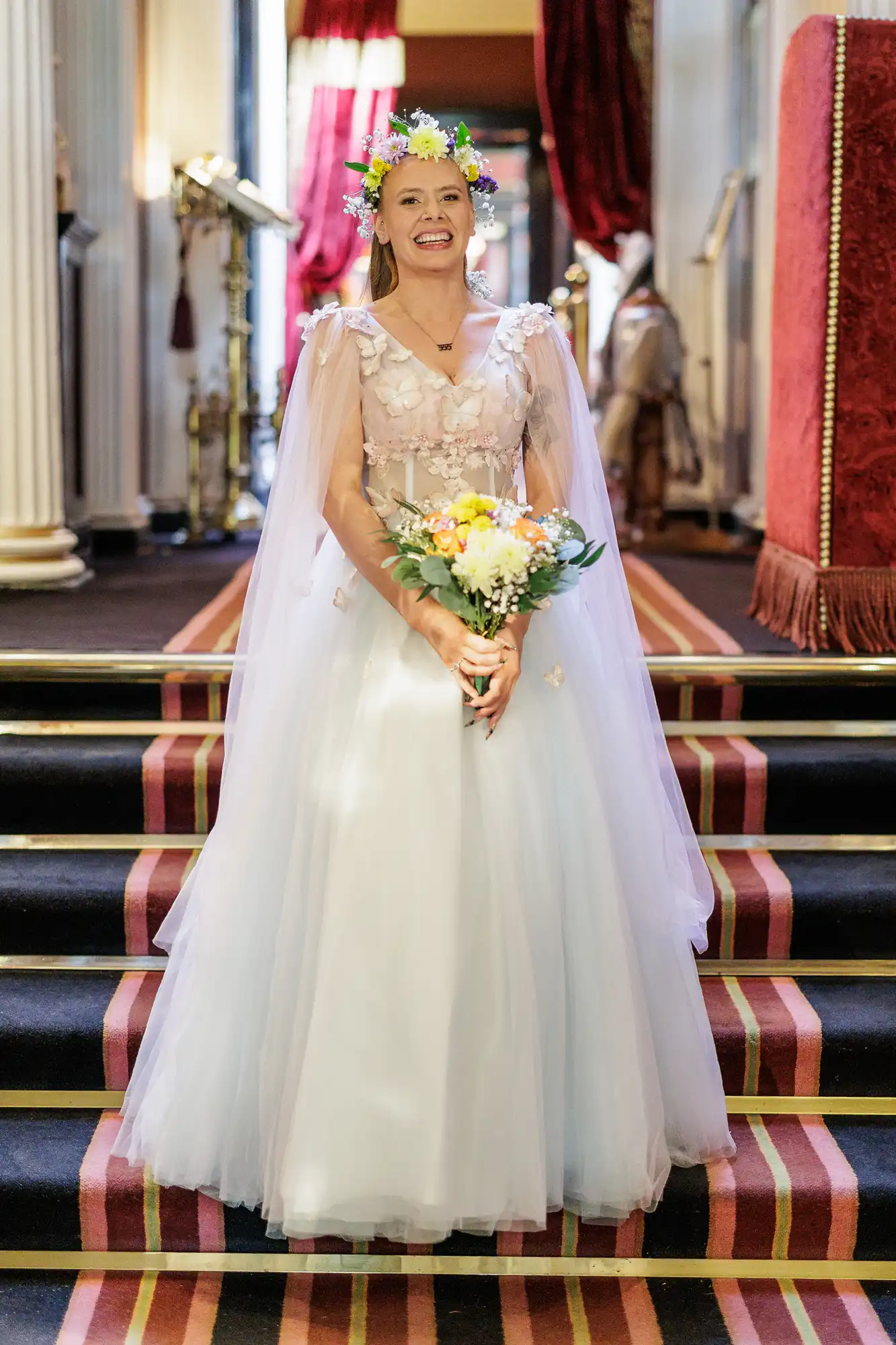 A smiling woman in a long, white tulle gown with a sheer, floral-embroidered cape stands on carpeted stairs. She wears a colourful flower crown and holds a bouquet of yellow and white flowers. Columns and ornate decor appear in the background.