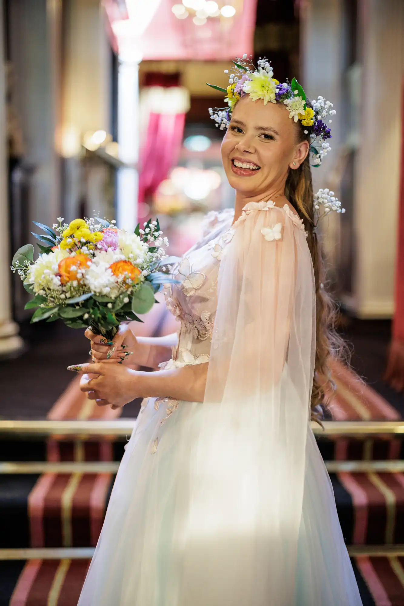 A smiling bride with long hair wears a pastel gown and a colourful flower crown. She holds a vibrant bouquet of flowers and stands indoors on a red carpet, with soft lighting and elegant décor visible in the blurred background.