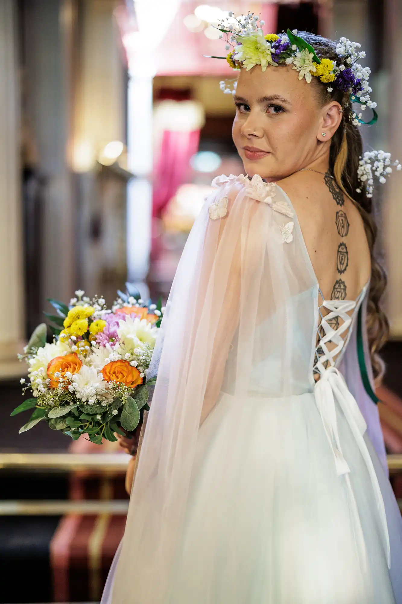 A bride in a white dress with sheer, floral-embellished sleeves looks over her shoulder. She wears a colourful flower crown and holds a bouquet of yellow, white, and orange flowers. A tattoo with geometric designs is visible on her back.