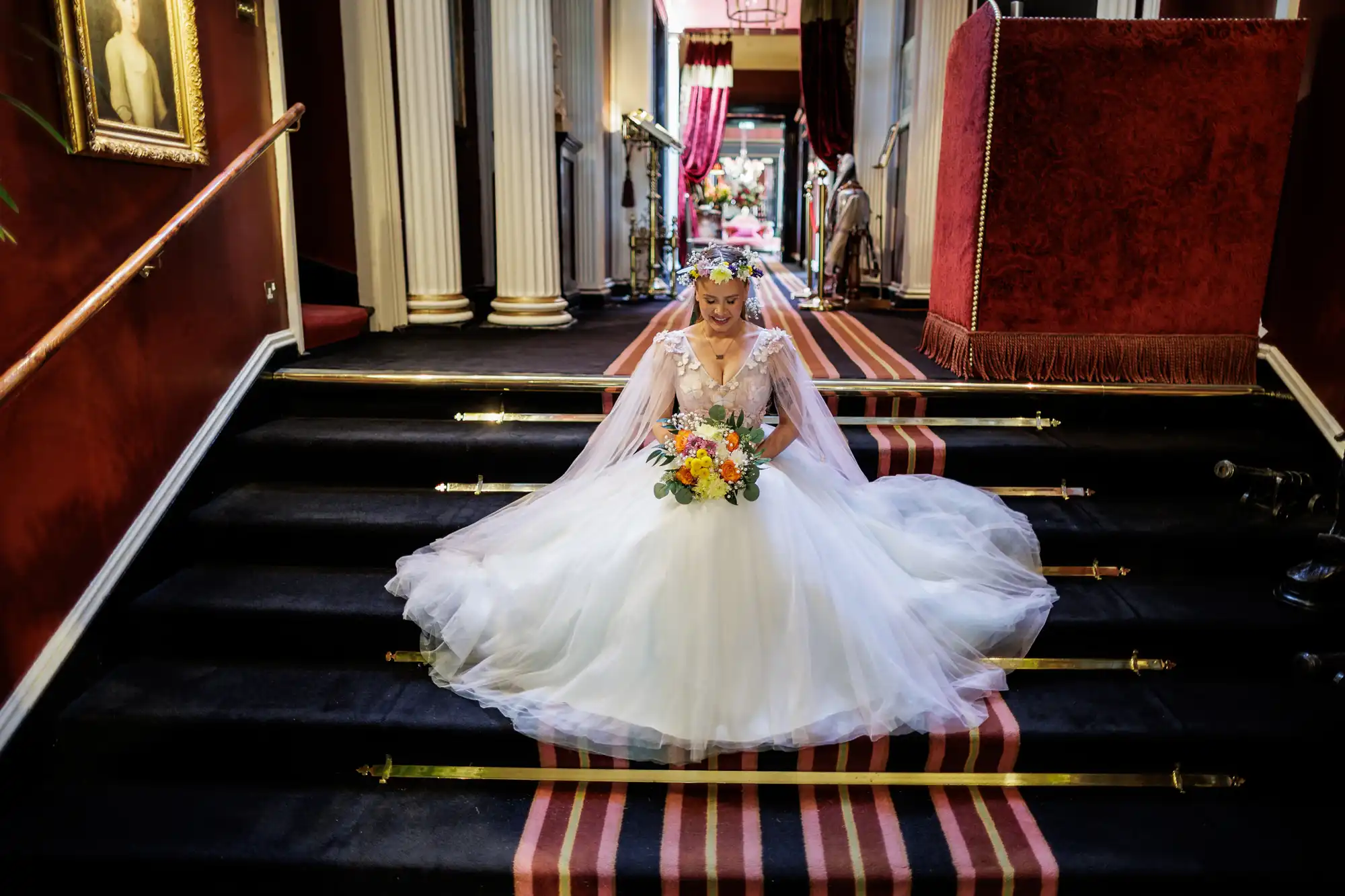 A bride in a full white gown with a long veil and flower crown sits gracefully on grand black-carpeted stairs. She holds a colourful bouquet and smiles softly. The elegant hallway features red walls, striped carpet, and tall white columns.