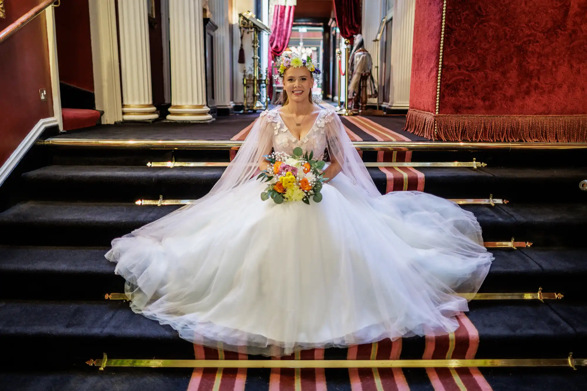 A bride in a white, full-skirted wedding dress with a floral crown and veil sits on black carpeted stairs, holding a colourful bouquet. Ornate red and gold decor, pillars, and draped curtains are visible in the elegant interior behind her.