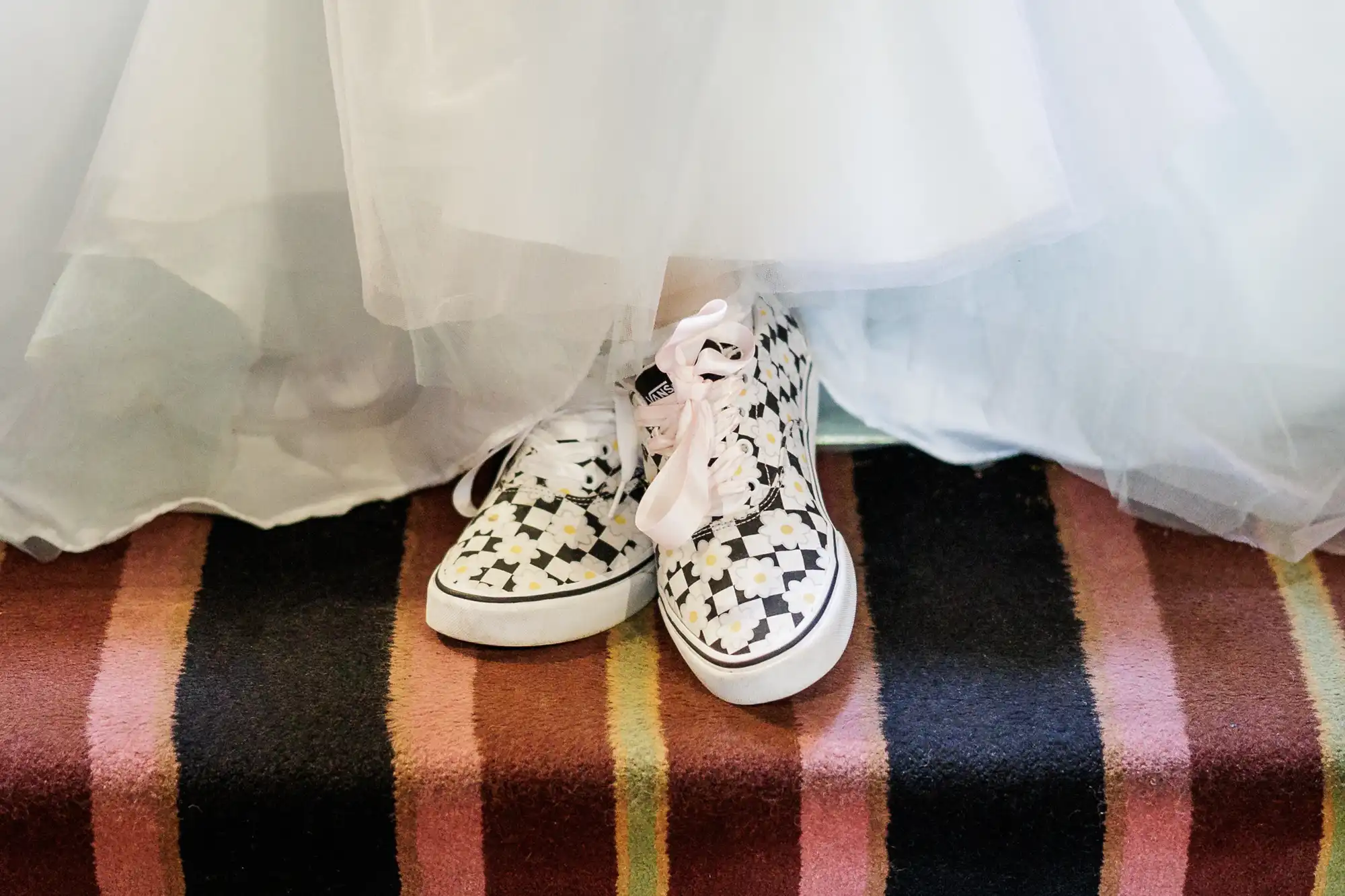 A person in a white tulle dress stands on a striped carpet, wearing patterned trainers with pink laces. Only the lower part of the dress and the shoes are visible, highlighting the contrast between formal attire and casual footwear.