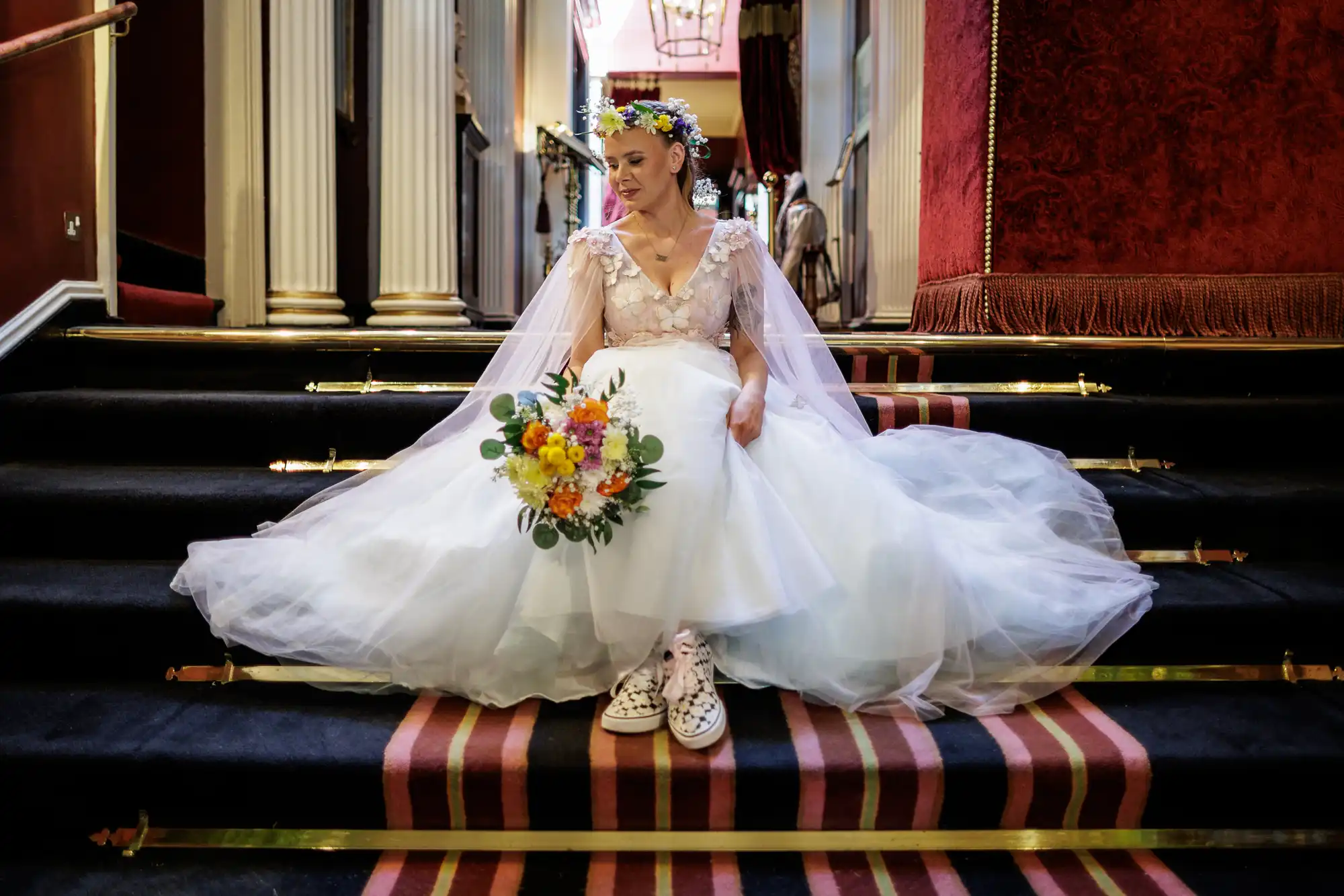 A bride in a white dress and floral crown sits on a staircase with red carpet and gold rails, holding a bouquet of yellow and orange flowers. She wears patterned trainers, and the setting features ornate pillars and deep red velvet walls.