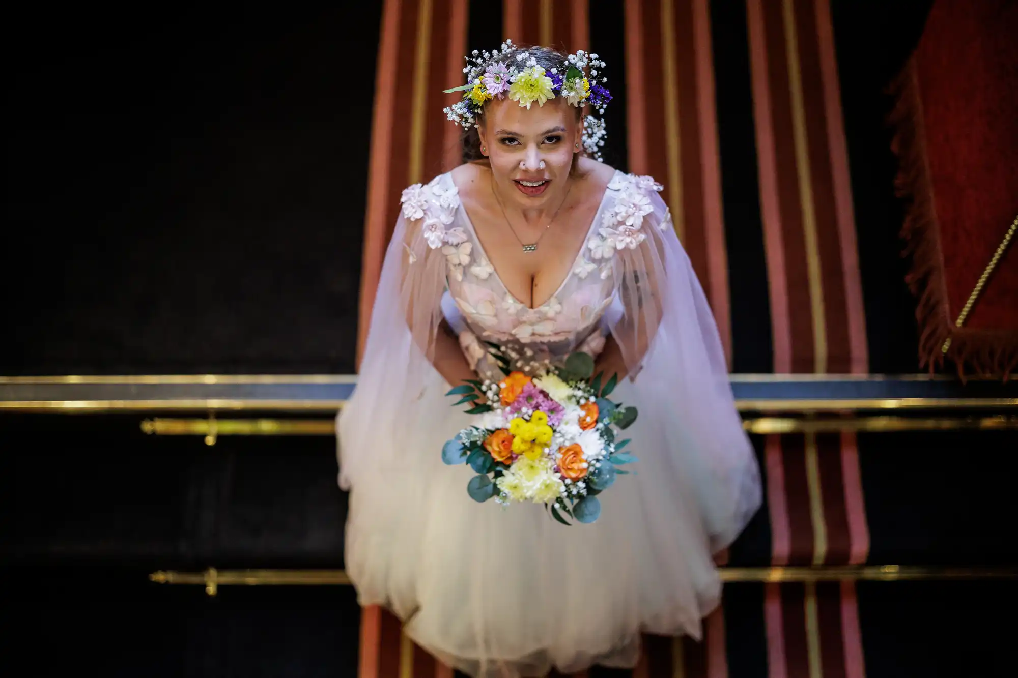A bride in a white dress with sheer sleeves and floral details looks up, smiling. She wears a colourful flower crown and holds a bouquet of orange, white, and yellow flowers. The photo is taken from above; red and gold carpeted steps are visible.