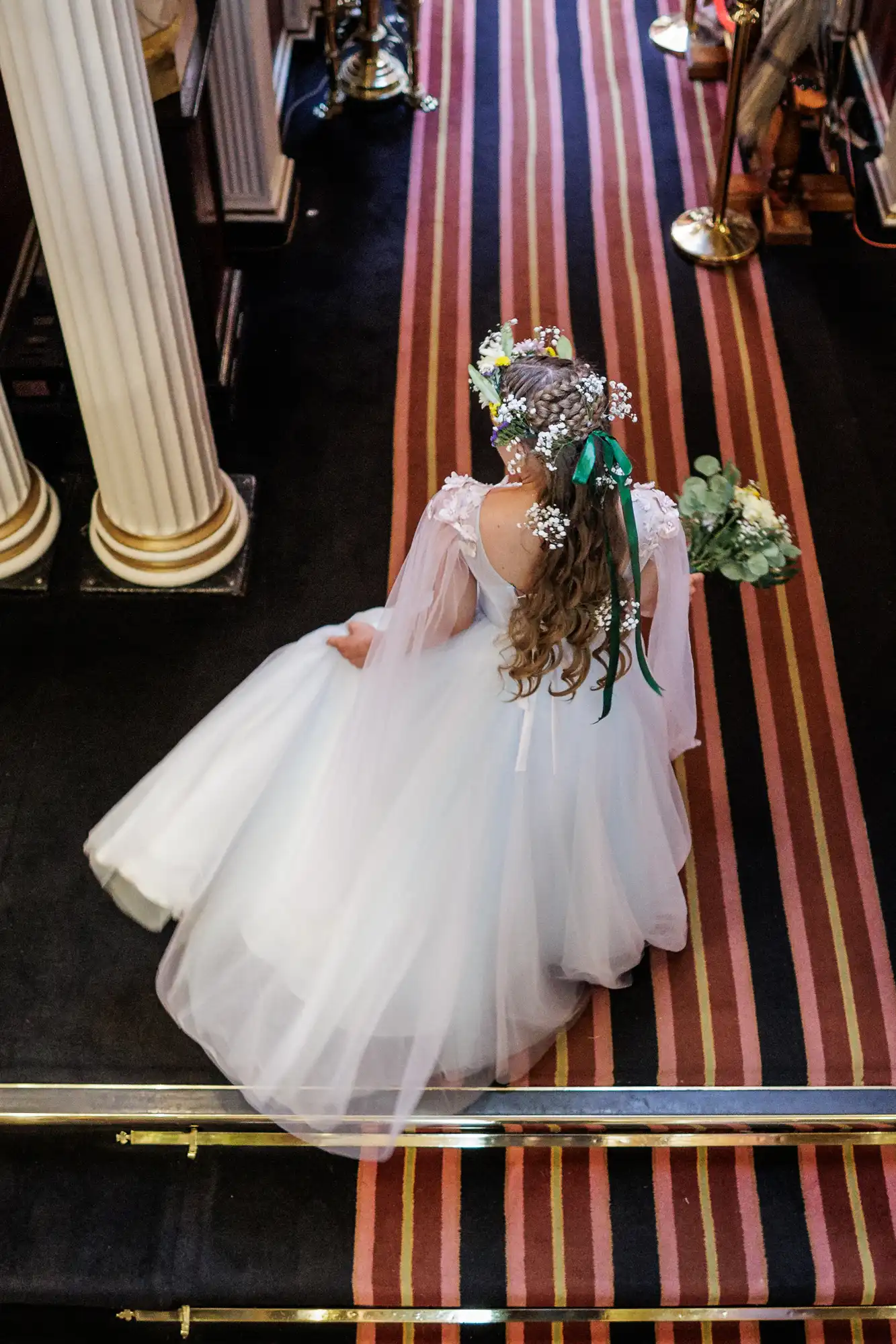 A bride in a white dress with flowing train and flower crown walks down a striped carpeted aisle in a formal indoor setting, seen from above. She holds a bouquet and her long hair cascades down her back, adorned with flowers and ribbon. White columns frame the scene.