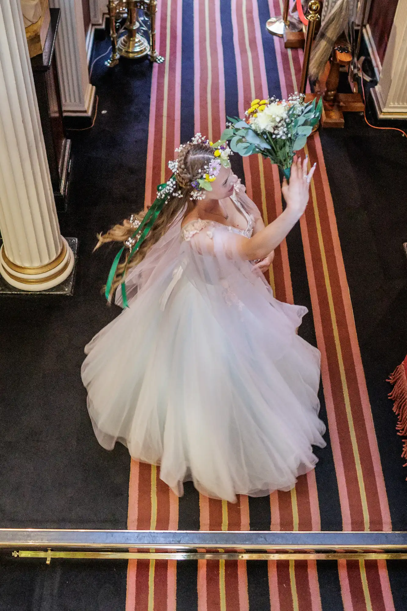 A bride in a white dress and floral crown stands on a striped carpet, holding a bouquet of yellow and white flowers aloft with both hands. Her hair is adorned with colourful flowers and ribbons. She is inside a decorated, columned venue, possibly a church.