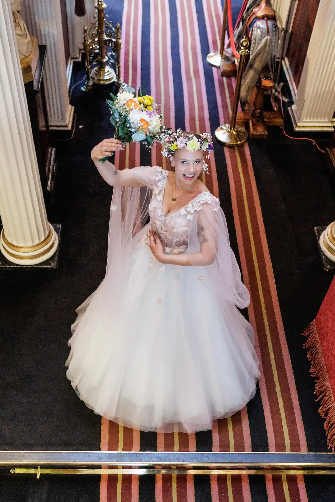 A smiling bride in a white gown and floral crown poses on a striped carpet between tall columns, holding a bouquet aloft and raising one arm gracefully, looking up towards the camera in a church or grand hall setting.