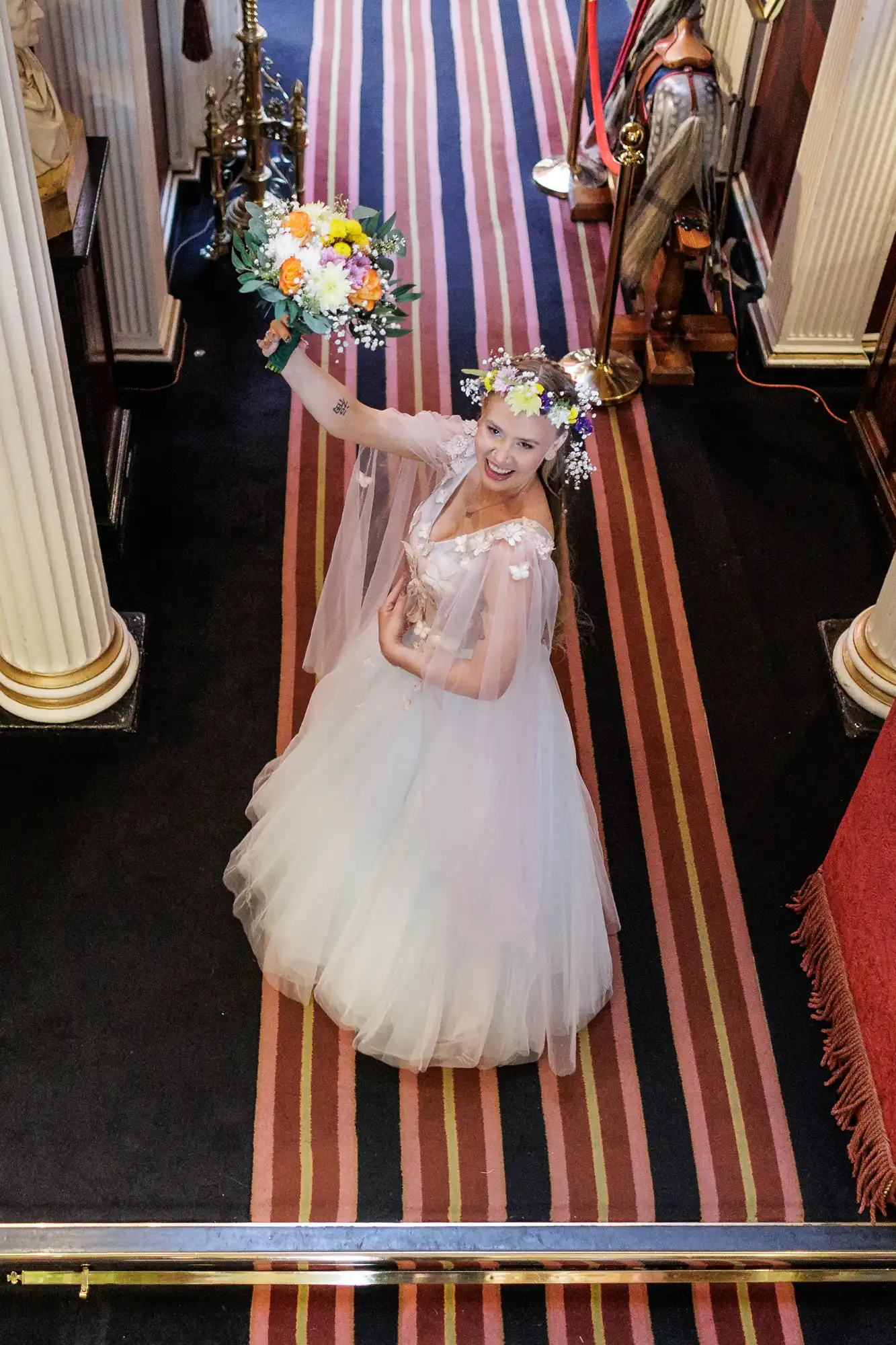 A smiling bride in a white gown and floral crown holds a bouquet of flowers aloft while standing on a striped carpet between white columns in a formal indoor setting. She looks up at the camera, surrounded by ornate décor.