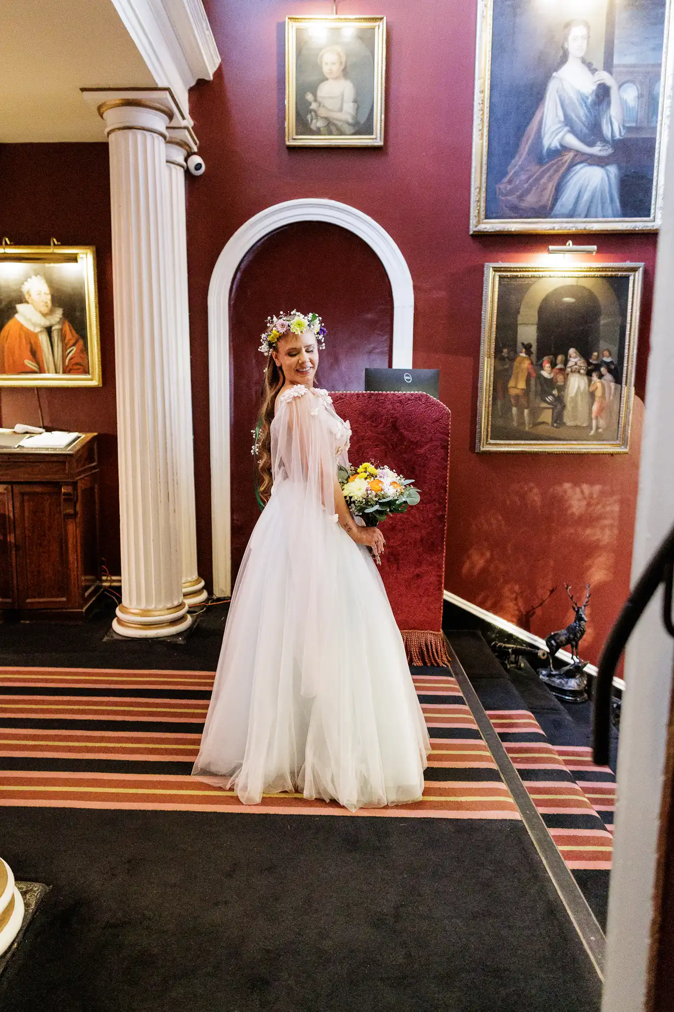 A smiling bride in a white dress and flower crown holds a bouquet, standing in a room with red walls, striped carpet, columns, and several framed portraits hanging on the wall behind her.