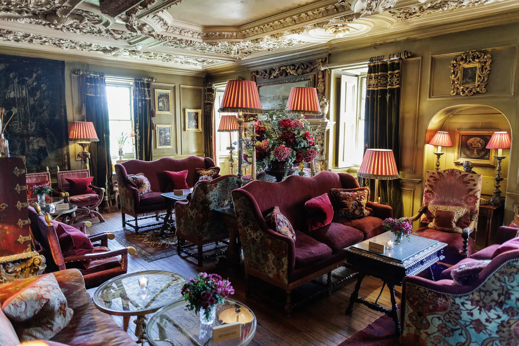 Ornate vintage living room with carved ceiling, tall windows, antique furniture, red velvet sofas, floral armchairs, patterned rugs, lamps with red shades, bouquets of flowers on tables, and classic artwork on the walls. Warm, cosy and richly decorated.