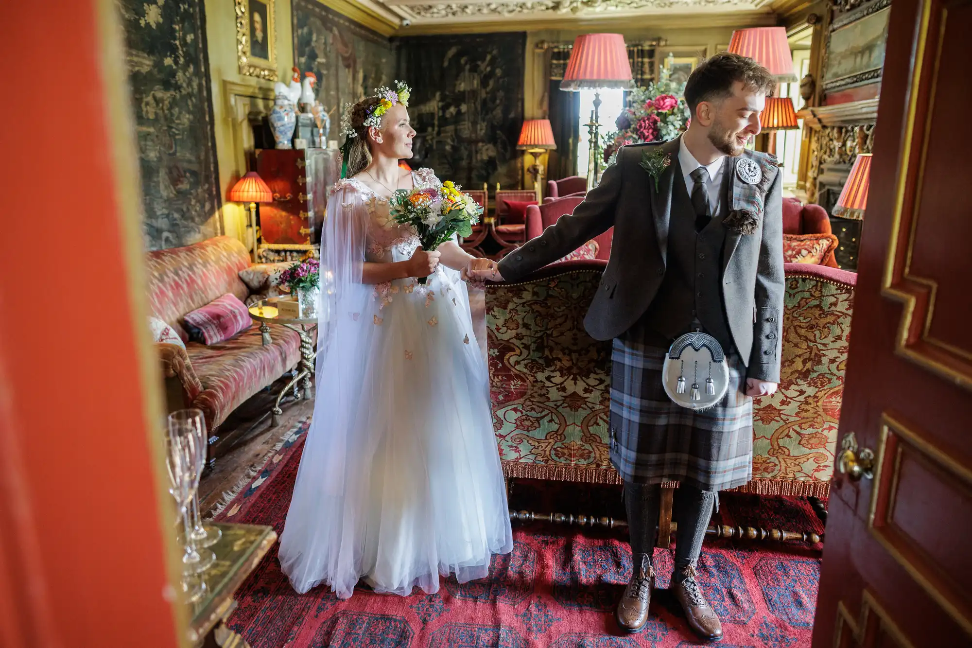 A bride in a white dress and floral crown holds a bouquet and smiles at a groom in a grey jacket, kilt, and sporran. They hold hands in an ornate room with antique furniture, red lamps, and framed art, standing on a red patterned rug.