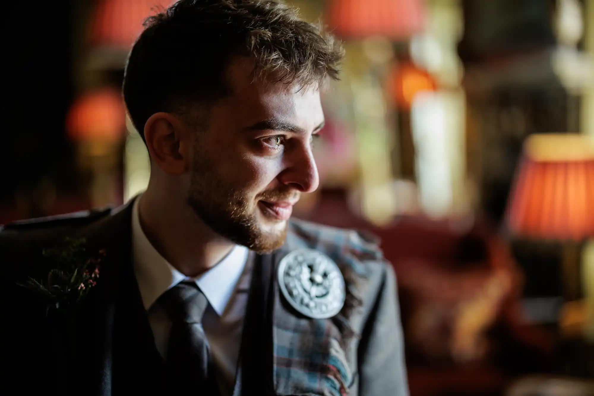 A man with short brown hair and a trimmed beard, wearing a grey checked jacket, white shirt, and tie with a decorative brooch, smiles whilst looking to the right; the background is softly blurred with warm, red-orange lighting from lamps.