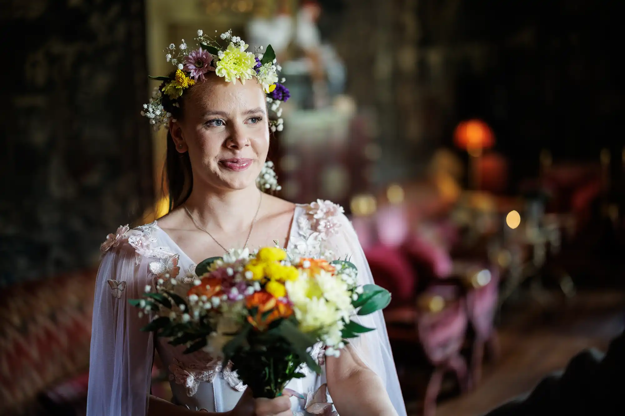 A woman wearing a white dress with floral details and a flower crown holds a colourful bouquet. She stands indoors in a softly lit, elegant room with blurred vintage furniture and warm lighting in the background, smiling gently.