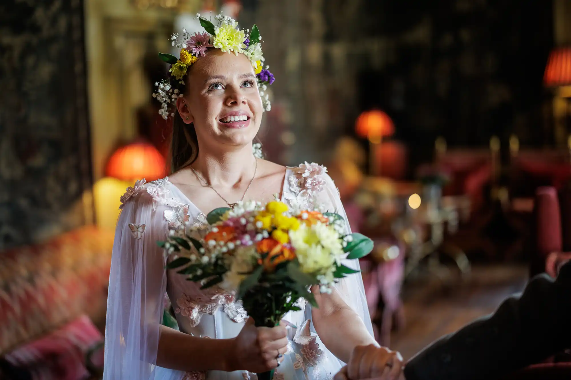 A smiling woman in a white floral dress and flower crown holds a colourful bouquet indoors. She looks upward in a warmly lit, elegant room with red lamps, patterned wallpaper, and plush seating in the background.