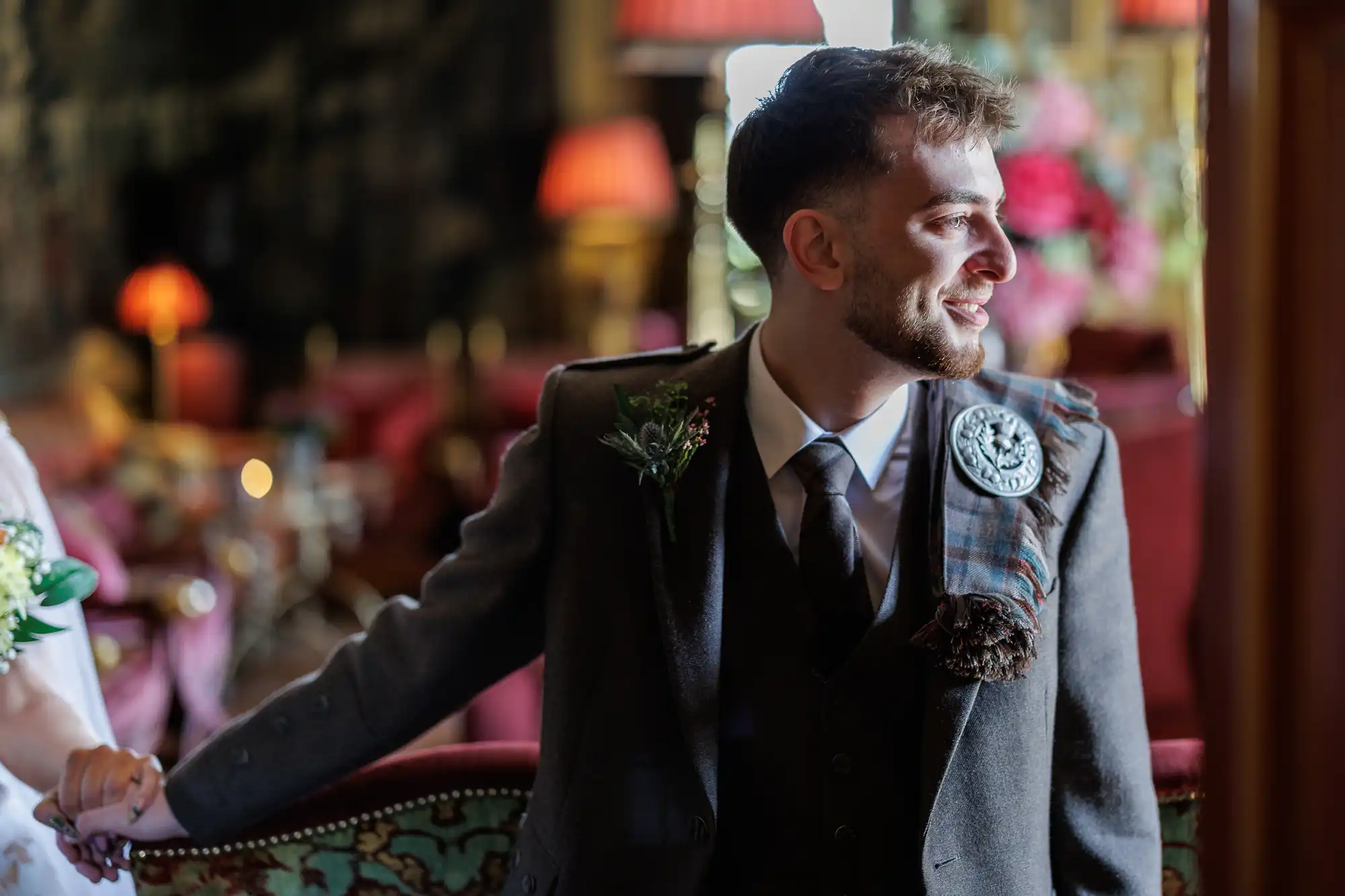 A man with short hair and a beard, wearing a dark suit, tie, and tartan sash with a decorative brooch, smiles whilst holding someone’s hand. He is seated indoors in a warmly lit, elegant room with blurred red lamps and flowers in the background.