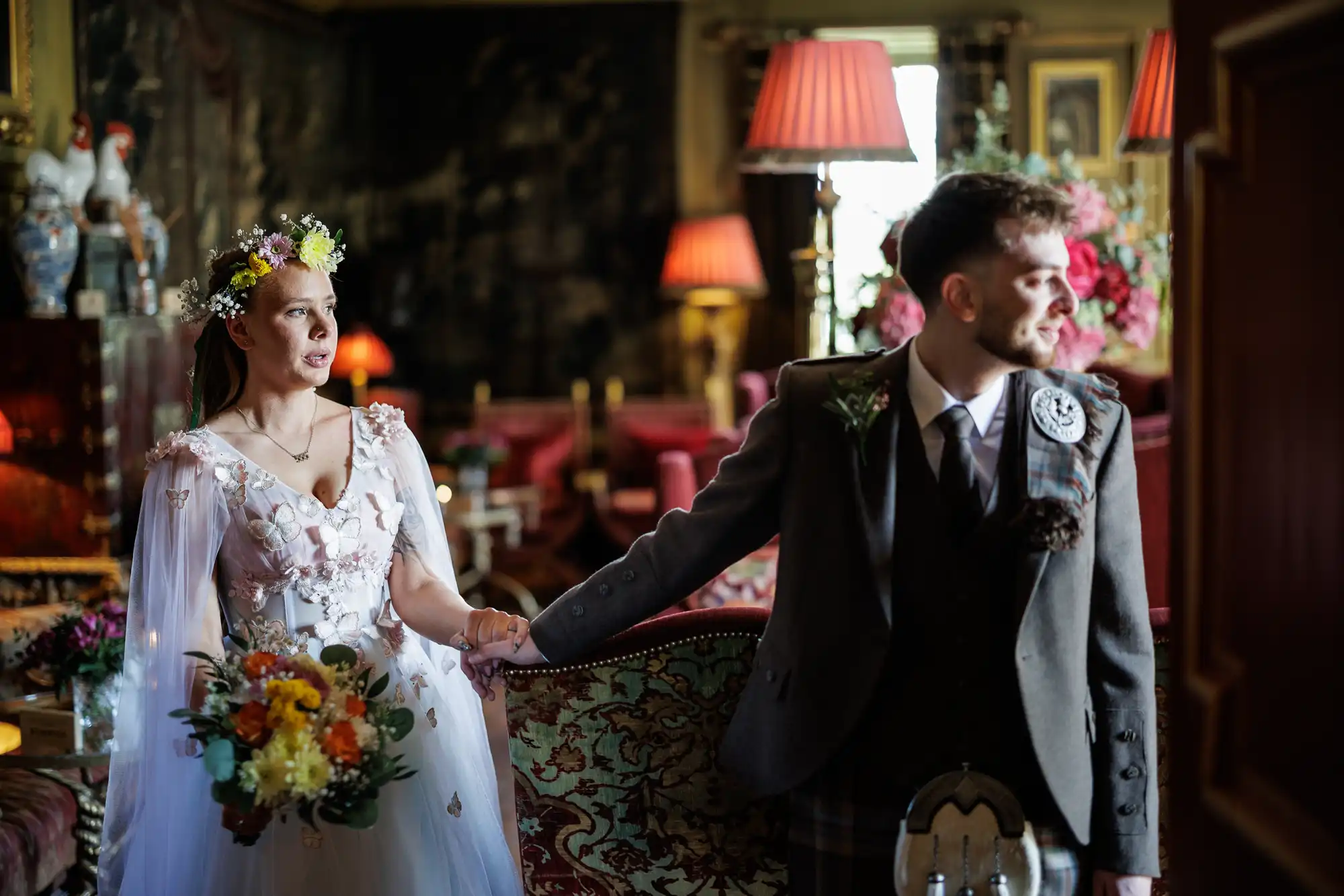 A bride in a white floral dress and flower crown holds a colourful bouquet and the hand of a groom in a kilt and jacket. They stand in an elegant, warmly lit room with red lamps, ornate decor, and floral arrangements. The groom looks away.