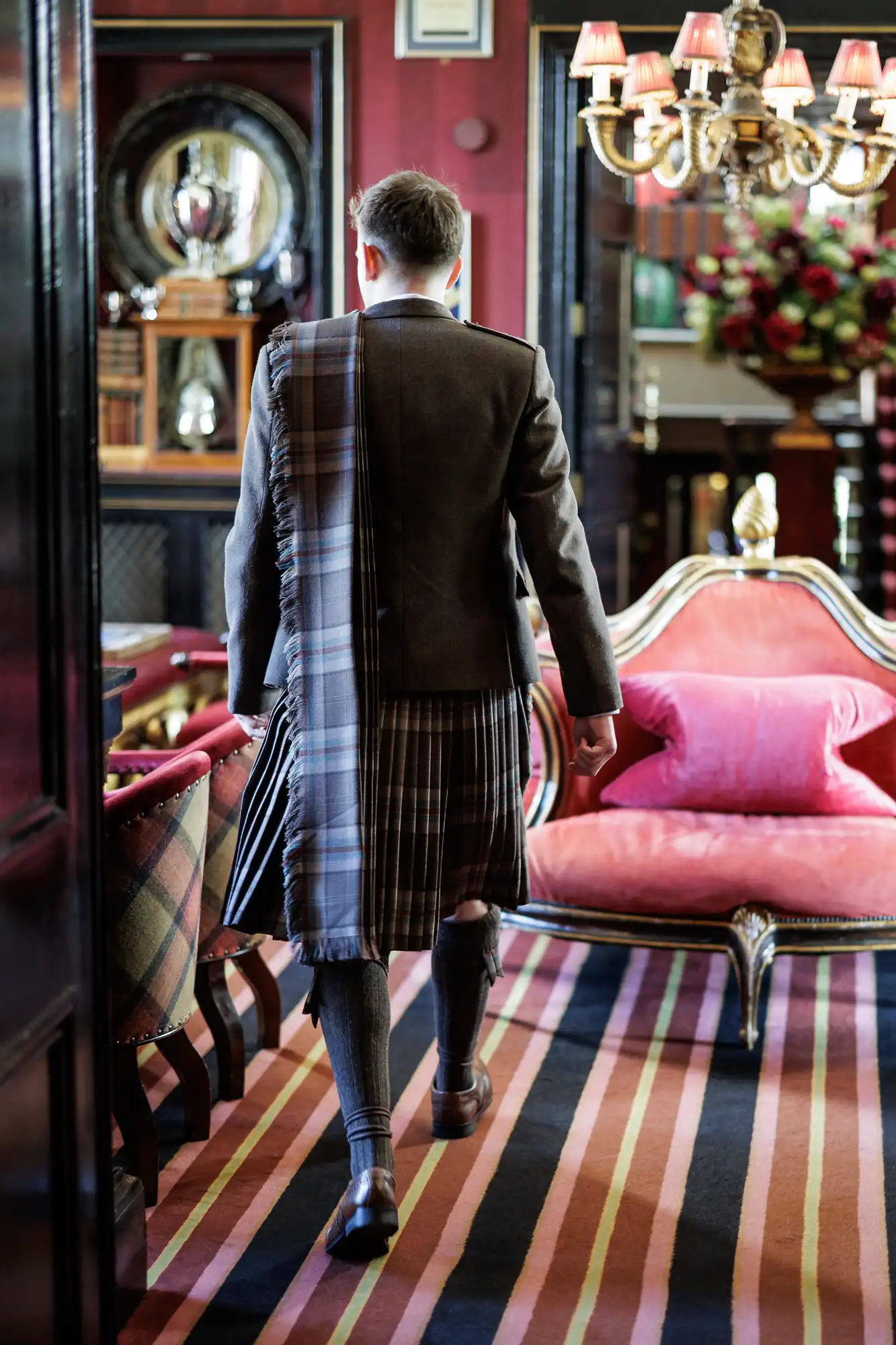 A man in a grey jacket and traditional Scottish kilt walks away in a lavish room with striped carpet, red velvet furniture, tartan accents, ornate decor, and a chandelier overhead. His attire features knee-high socks and a long tartan sash.