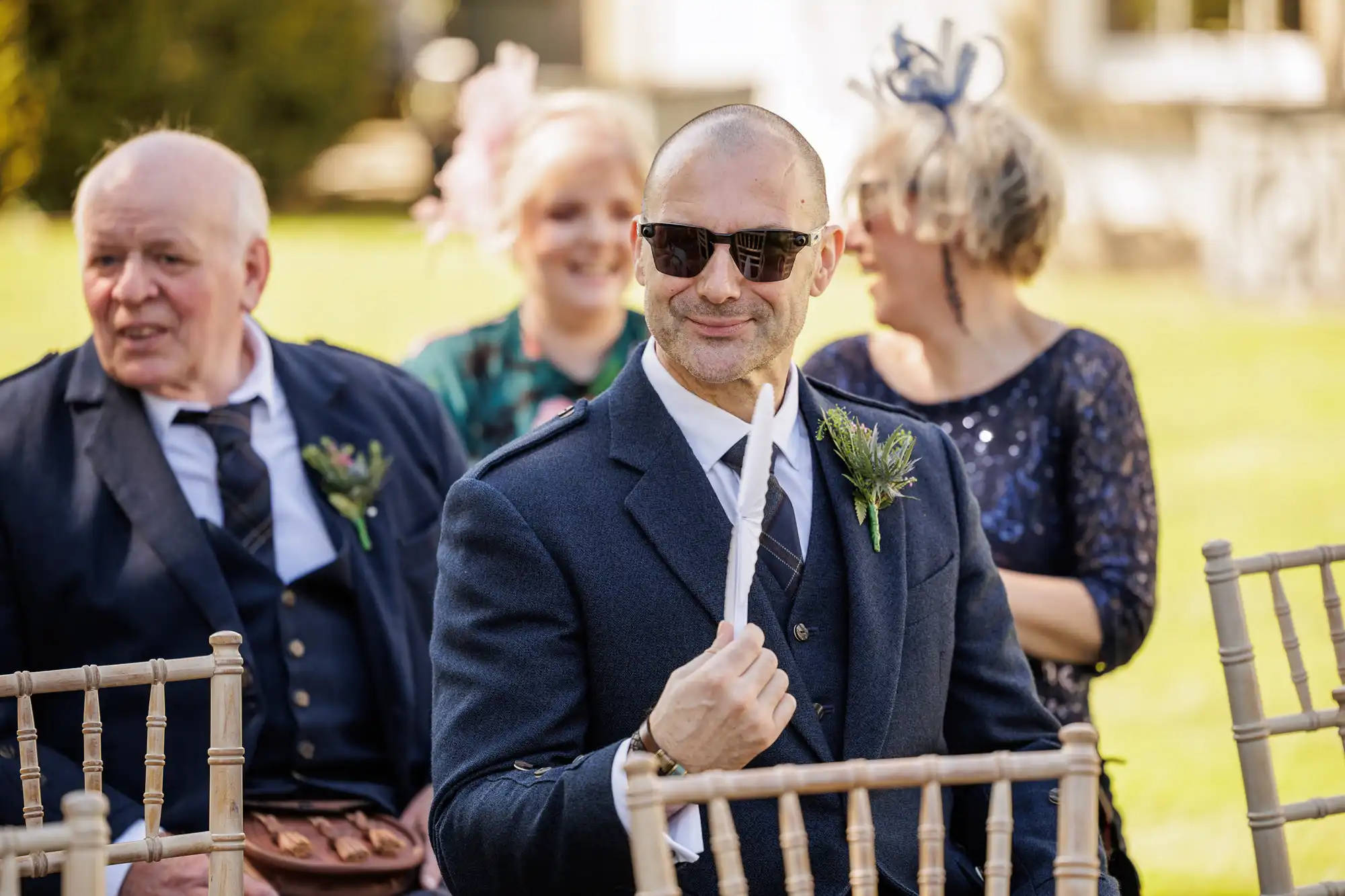 A man in a dark suit, sunglasses, and a thistle buttonhole sits outdoors on a wooden chair, smiling and holding a white paper. Other people, dressed formally, sit around him, with greenery and sunlight in the background.
