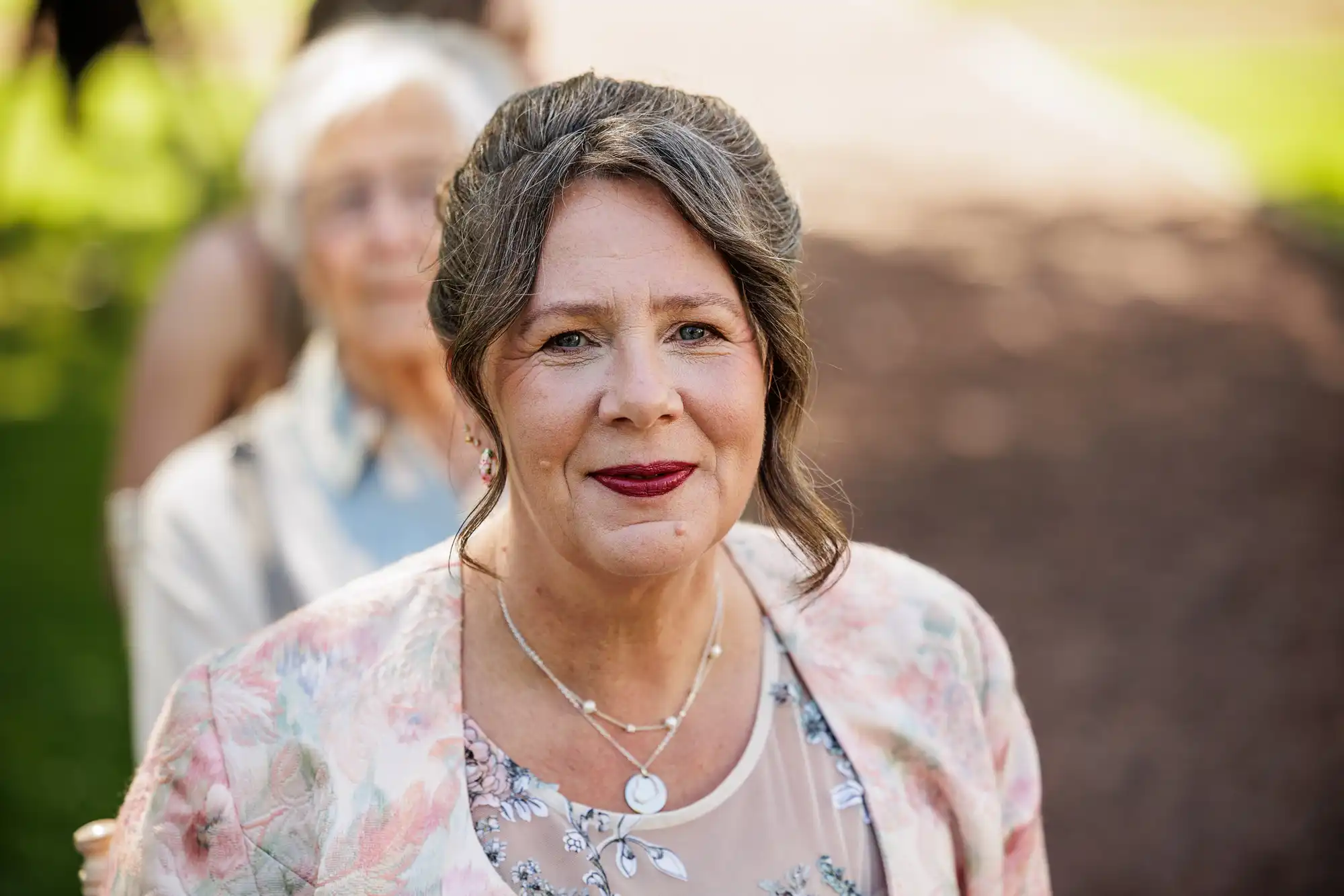 A middle-aged woman with brown hair styled up and wearing a floral jacket over a sheer top smiles gently at the camera outdoors. She has dark red lipstick and layered necklaces. An older woman with white hair is blurred in the background.