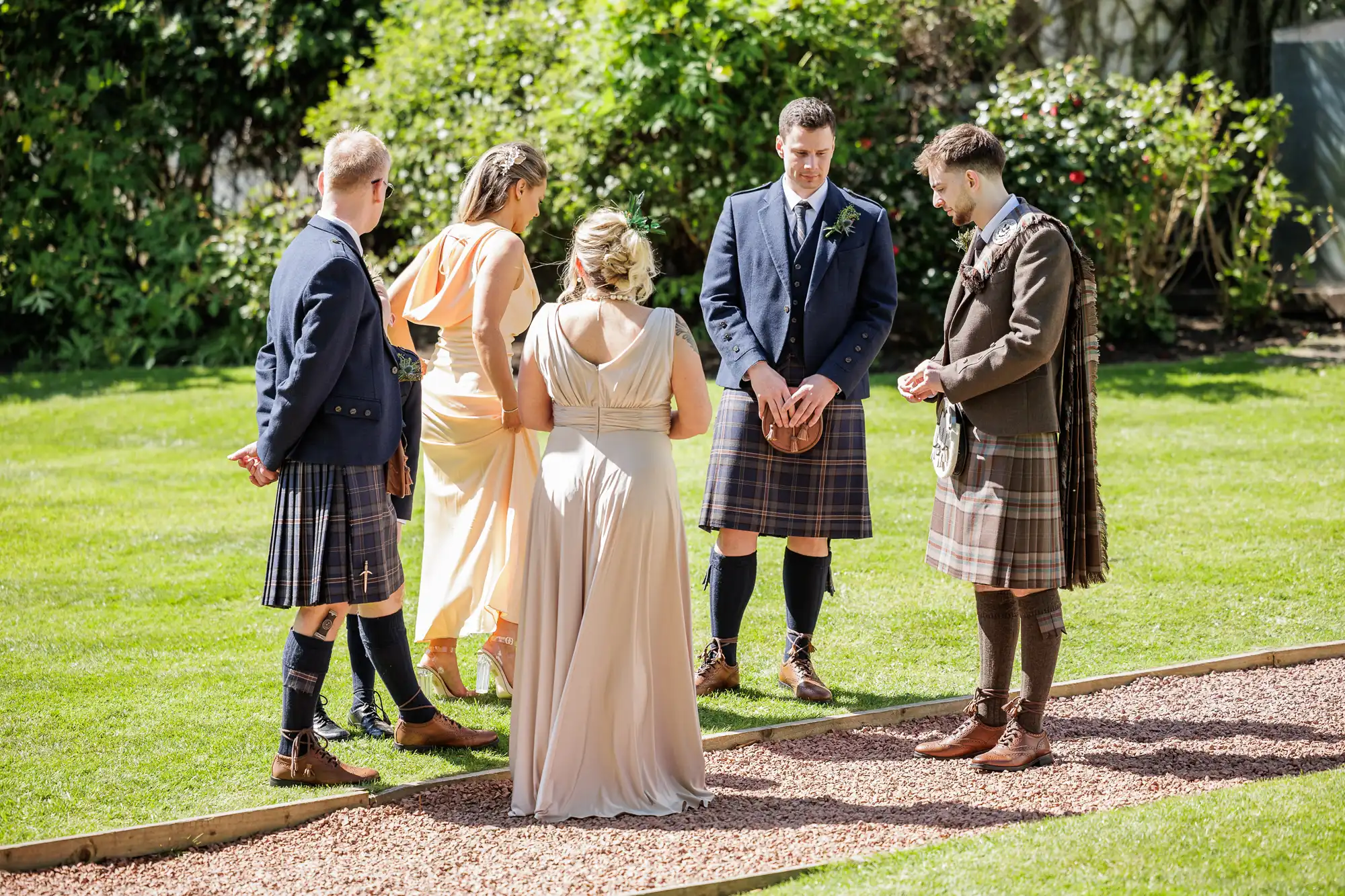 Five people stand on a sunlit lawn, four wearing kilts and one in a peach dress. The women face away from the camera, while the three men and one woman converse, surrounded by greenery and dressed in formal attire, suggesting a special outdoor event.