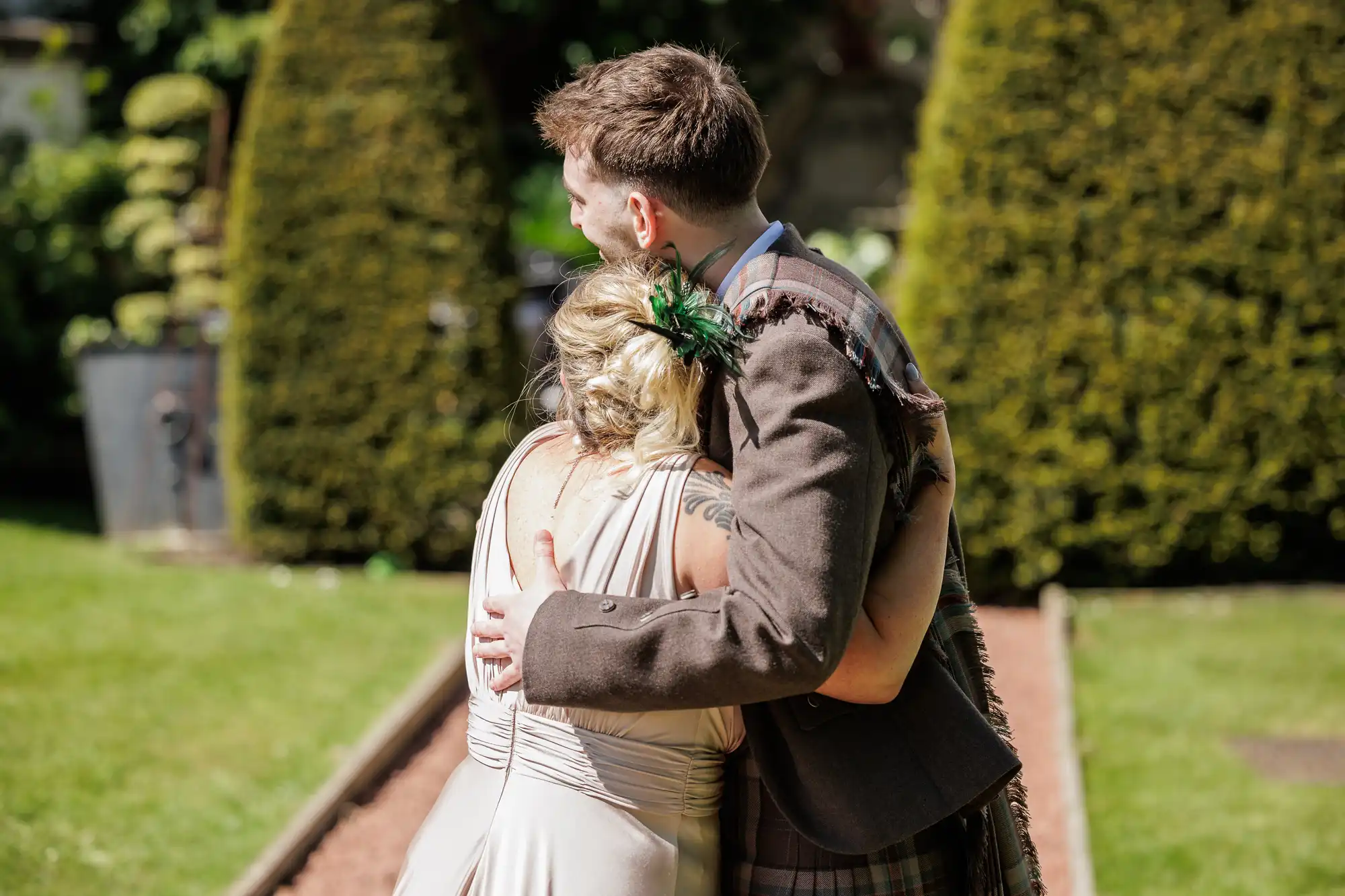 A man in a brown jacket and checked kilt hugs a woman with blonde hair in a bun and a pale dress. They stand outdoors on a sunny day with trimmed hedges and grass behind them. The woman has a feather hairpiece and a visible tattoo on her shoulder.
