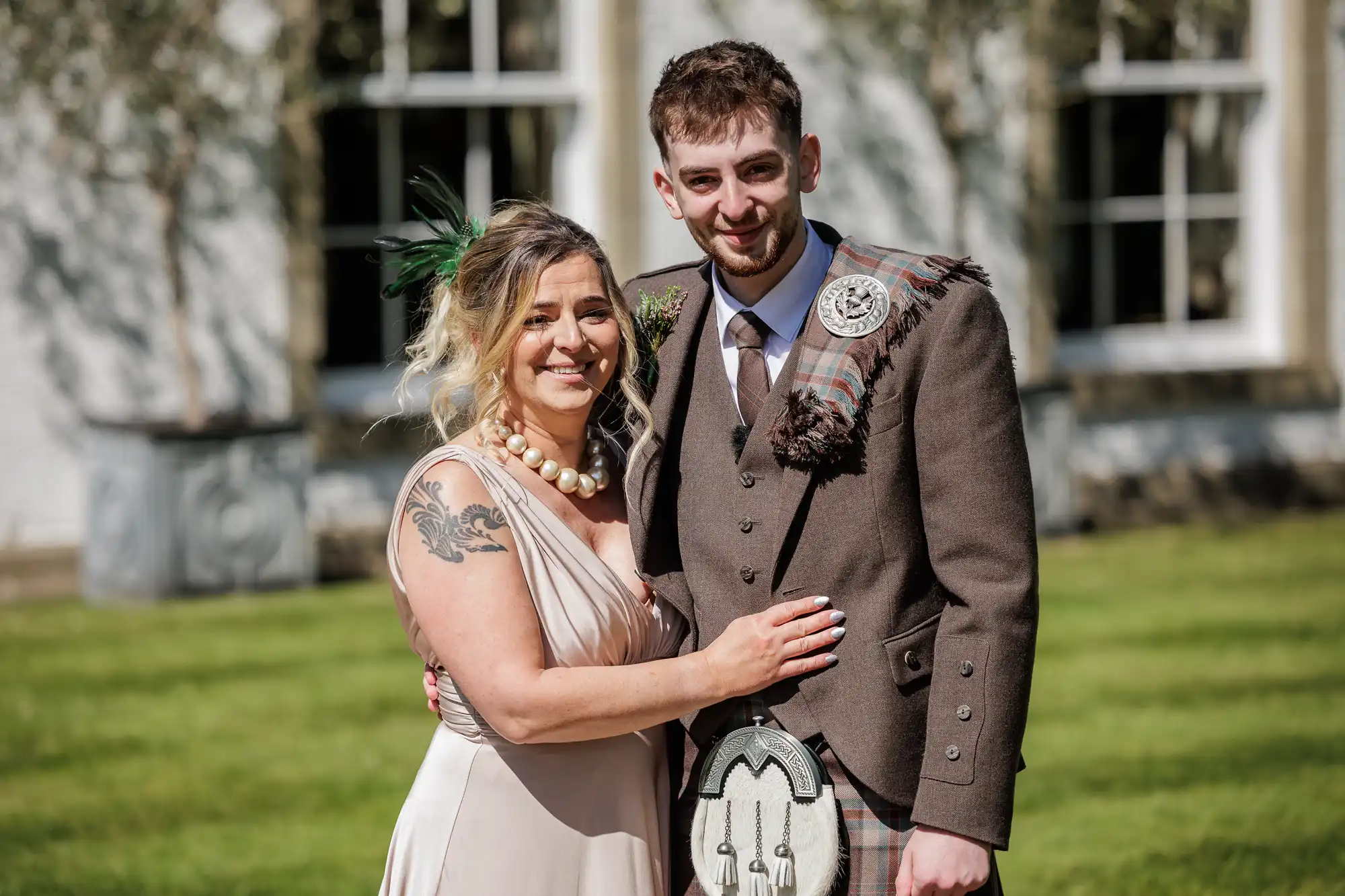 A smiling woman in a pale pink dress with a large pearl necklace and arm tattoo stands next to a man in traditional Scottish attire, including a brown jacket, kilt, and tartan sash. They stand outside on a lawn with a building and windows behind them.