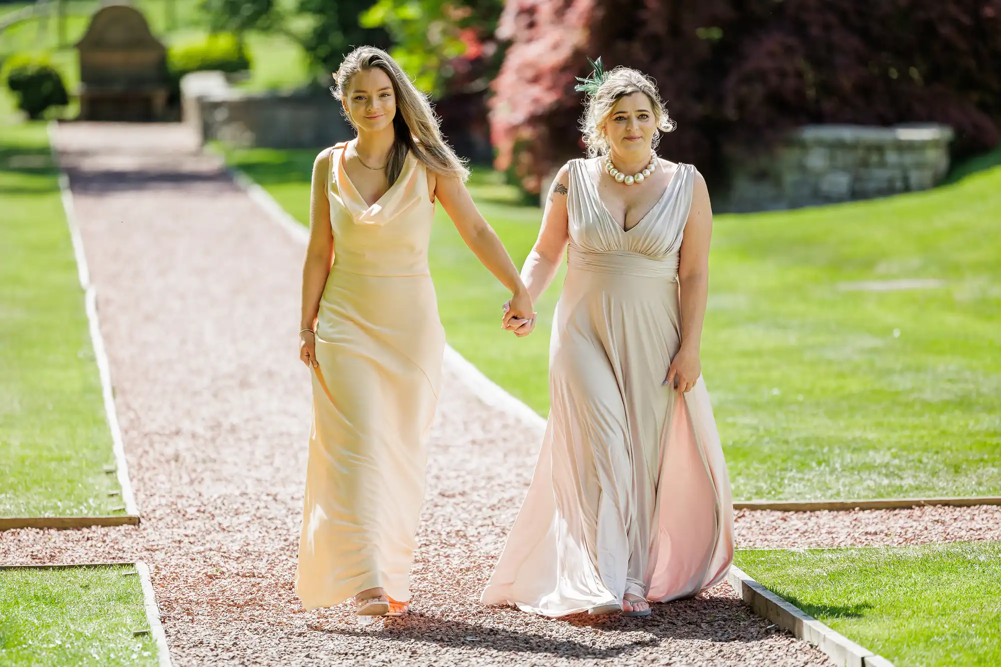 Two women in pale, flowing dresses walk hand-in-hand on a sunlit garden path. Both have long hair, and one wears a floral headpiece and necklace. Green grass and trees surround them, with sunlight highlighting the scene.