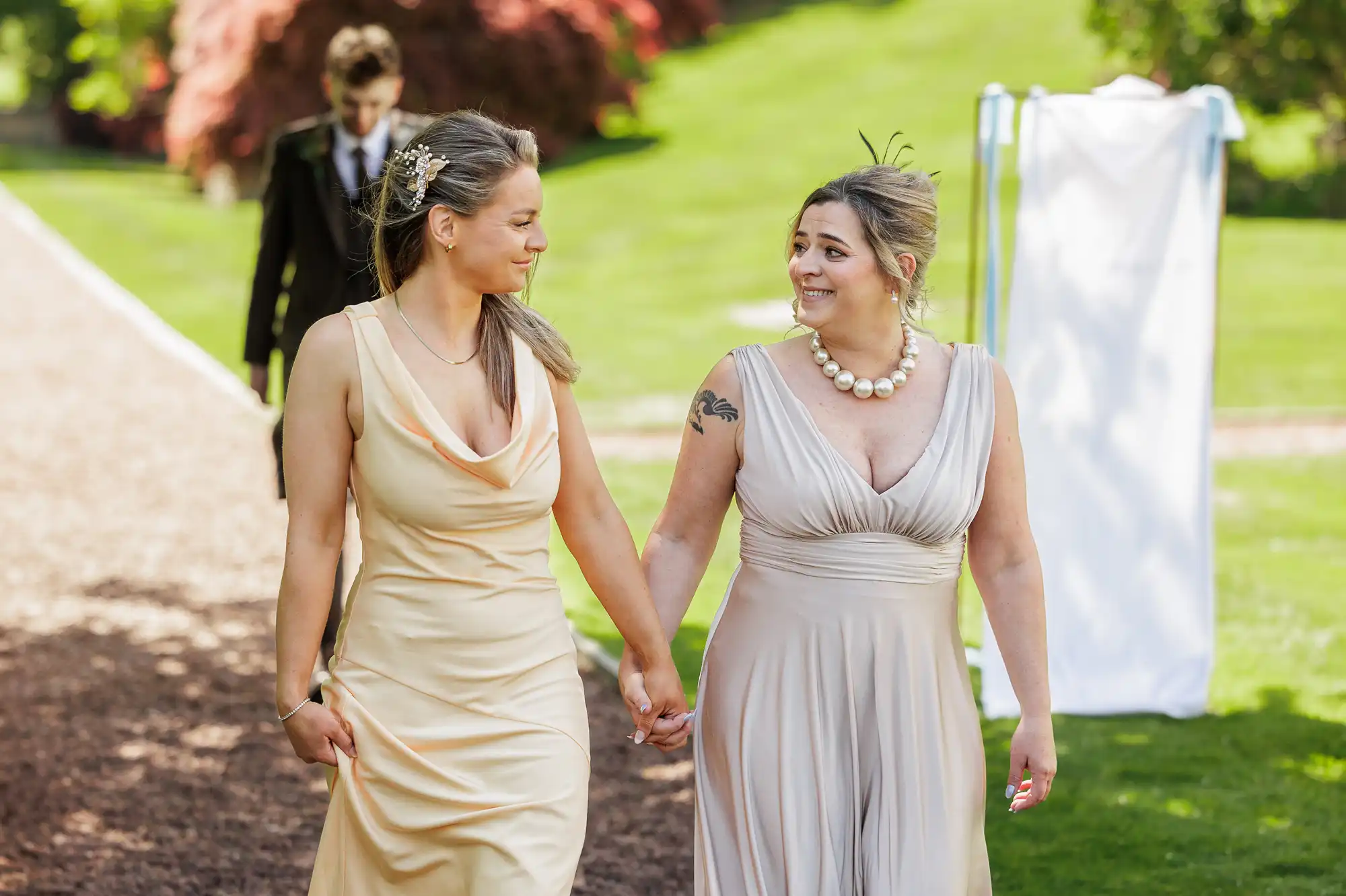 Two women in light-coloured dresses walk hand in hand outdoors, smiling at each other. One wears a pearl necklace and has a visible tattoo on her arm; a man in a suit walks behind them on a sunny, green lawn with trees and a white draped structure.